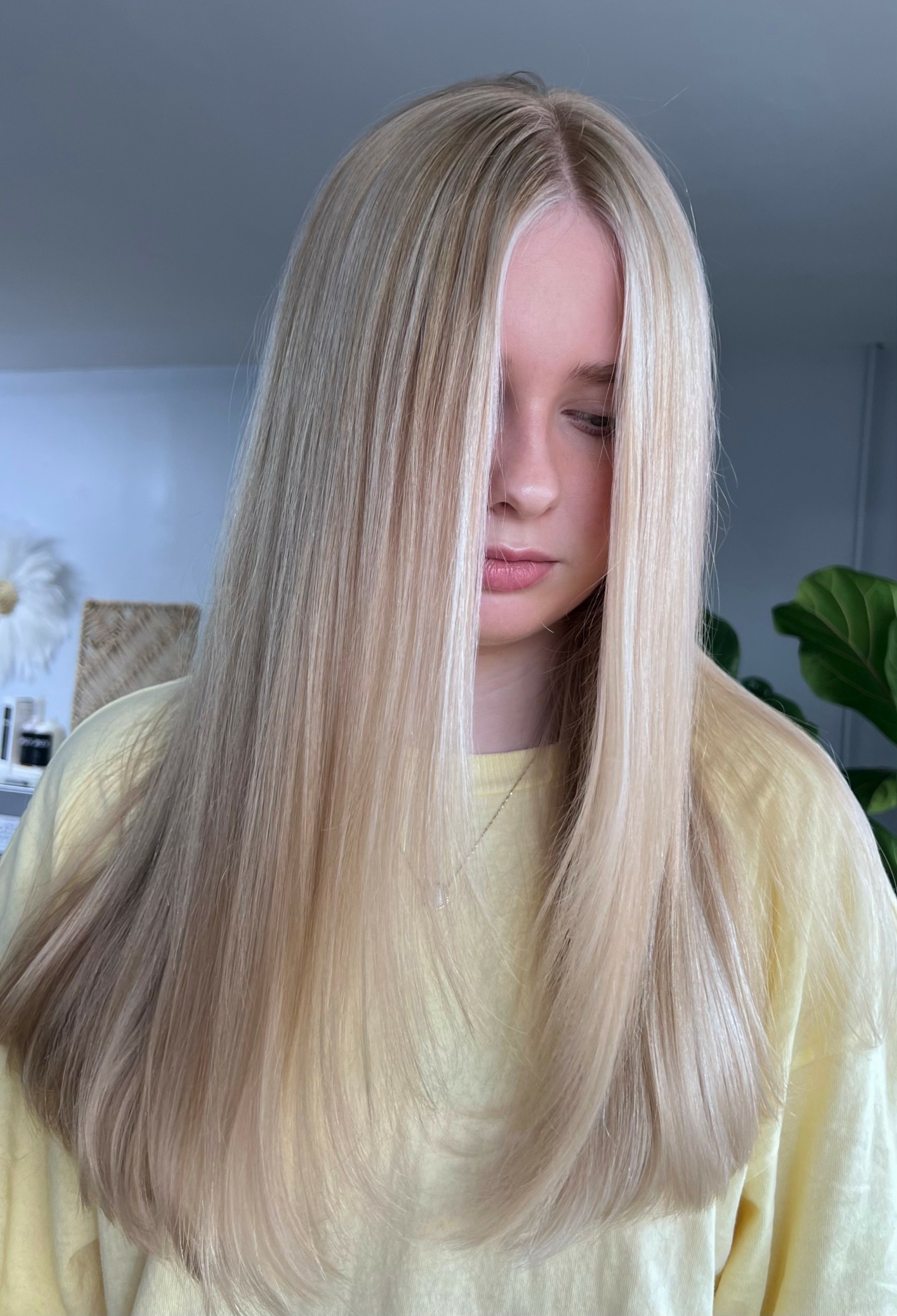 Young woman with long, straight blonde hair and closed eyes, wearing a light yellow top, indoors with plants in background.