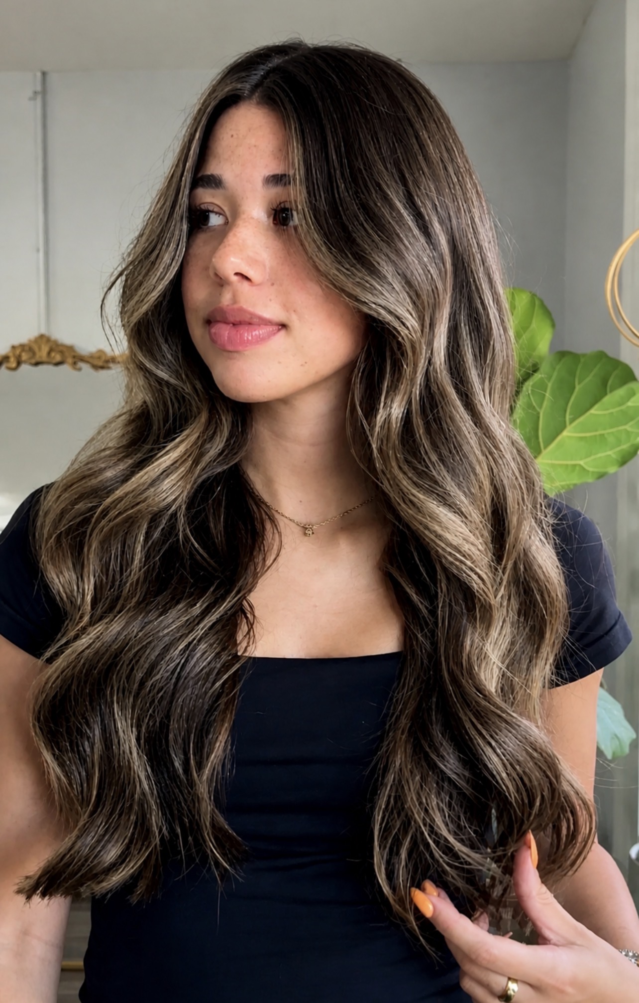 Woman with long wavy hair and a black top, standing indoors near a green plant.