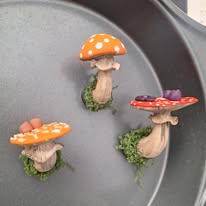 Three small mushrooms with orange, red, and purple caps on a gray surface, with green moss at their bases.