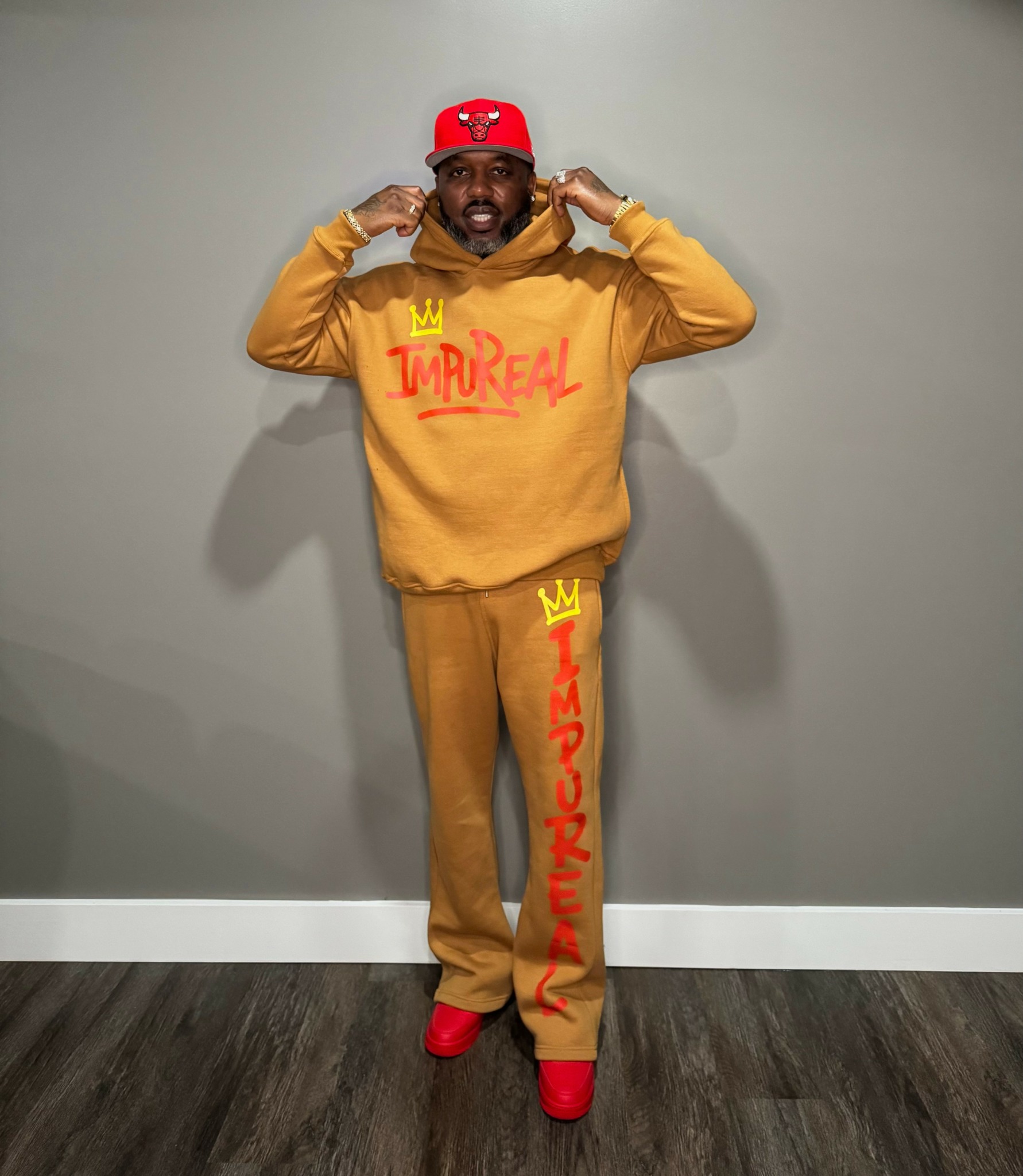 Man in yellow hoodie and pants with red text, red cap, and red shoes, standing against a gray wall.