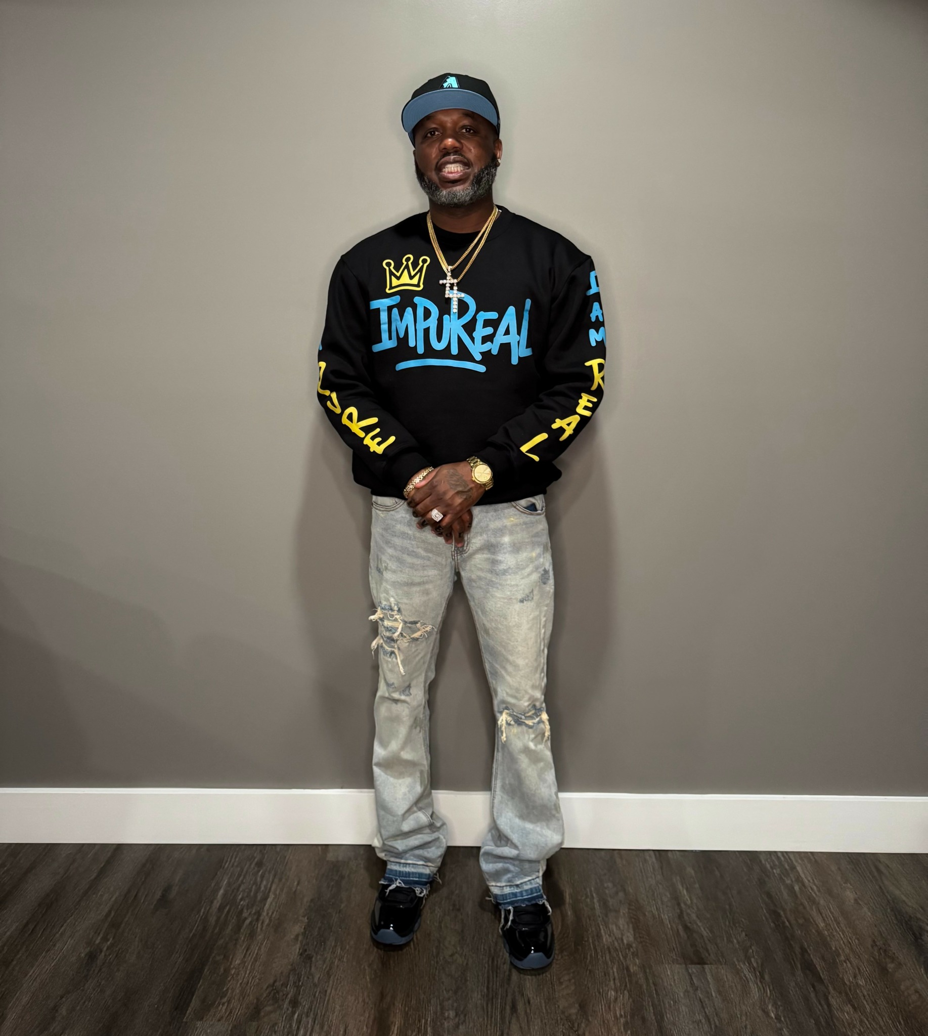 Man standing against a plain wall, wearing a black sweatshirt with colorful text and graphics, light jeans, and sneakers.