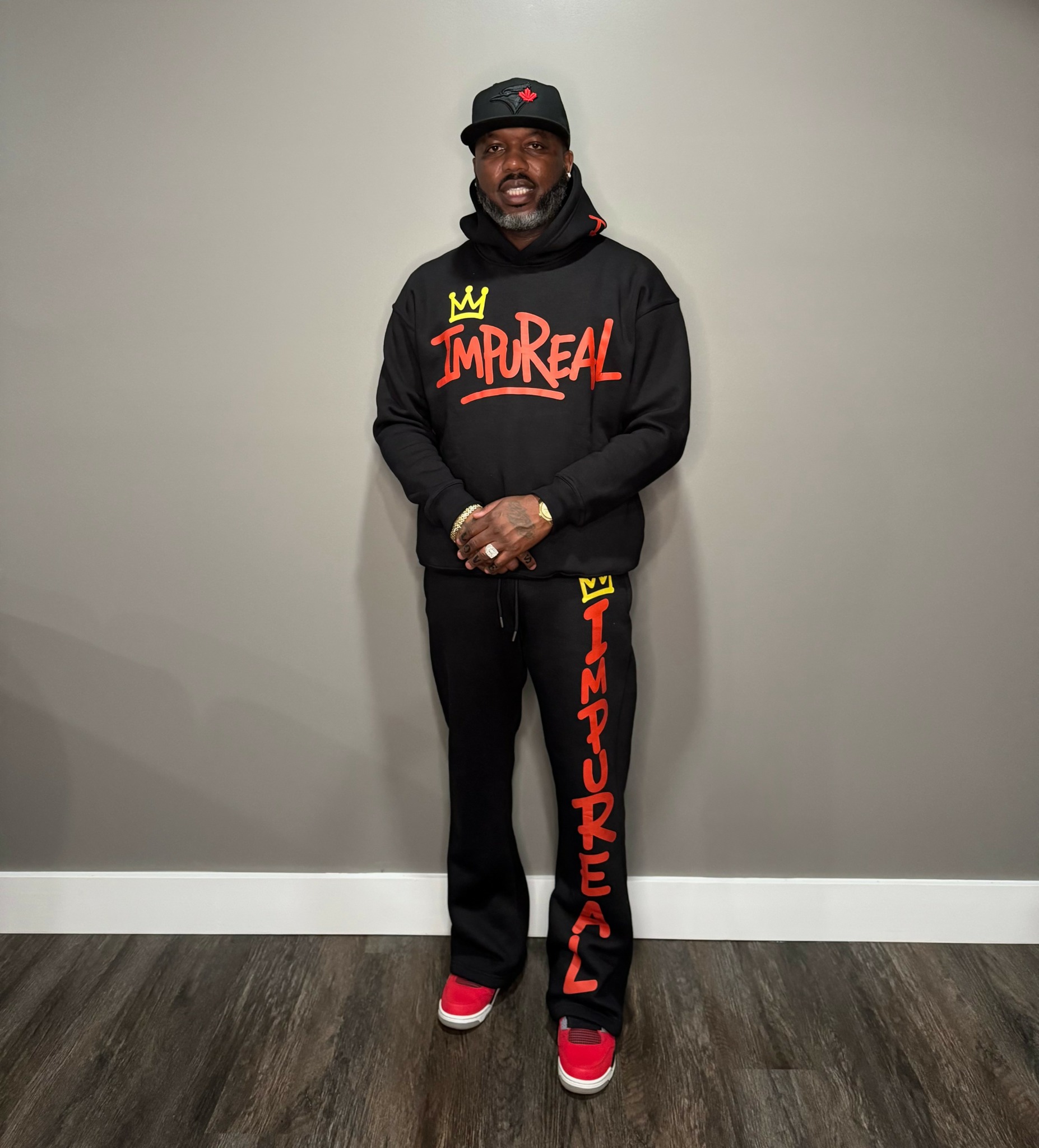 Man standing against a plain wall, wearing a black hoodie and pants with red and yellow text, red shoes, and a black cap.