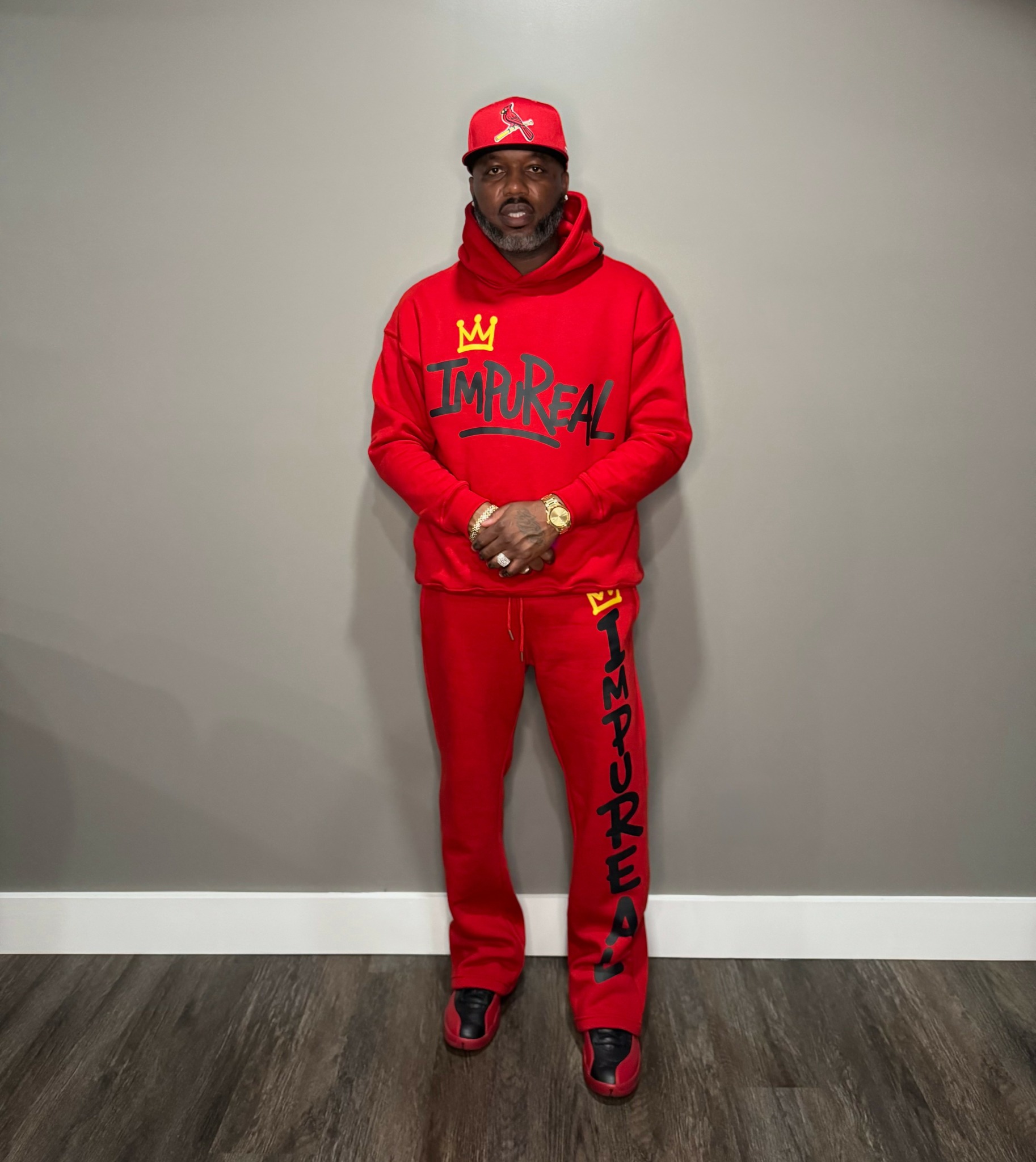Man in red hoodie, sweatpants, and cap stands against gray wall, hands clasped, looking at camera.