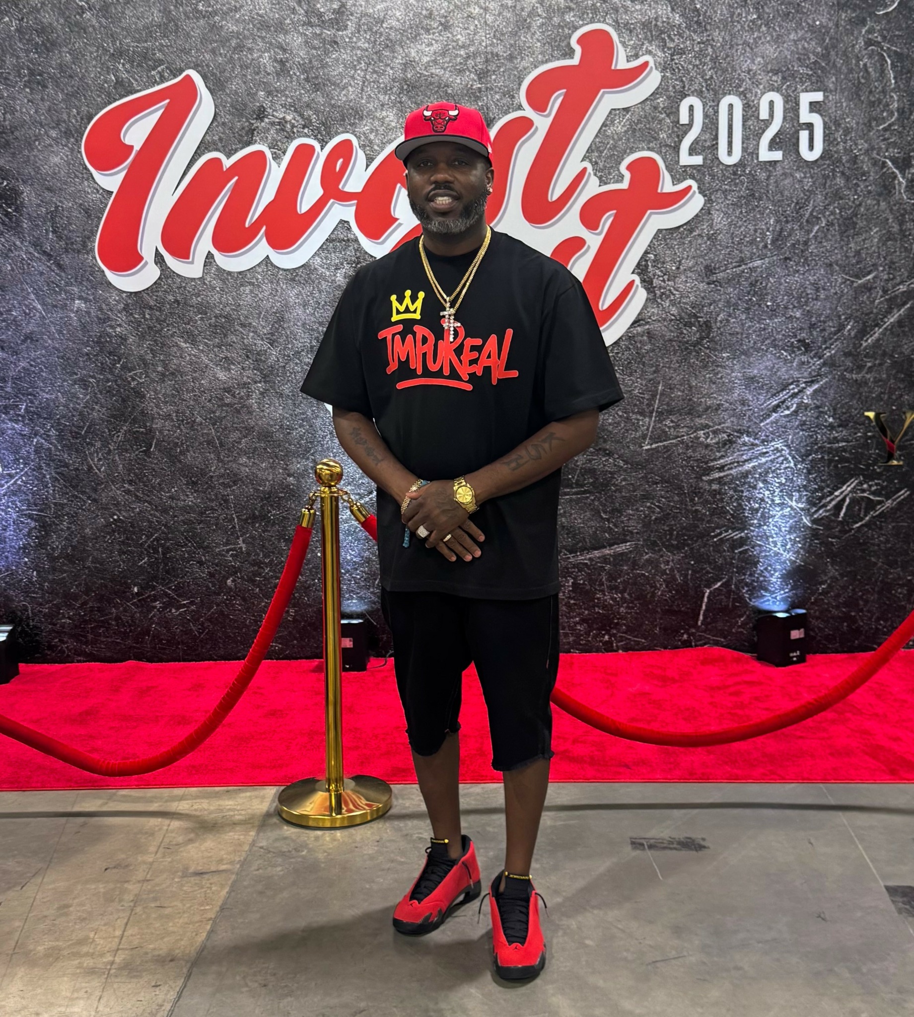Man standing in front of a backdrop with red carpet and gold stanchions, wearing a black T-shirt, shorts, and red sneakers.