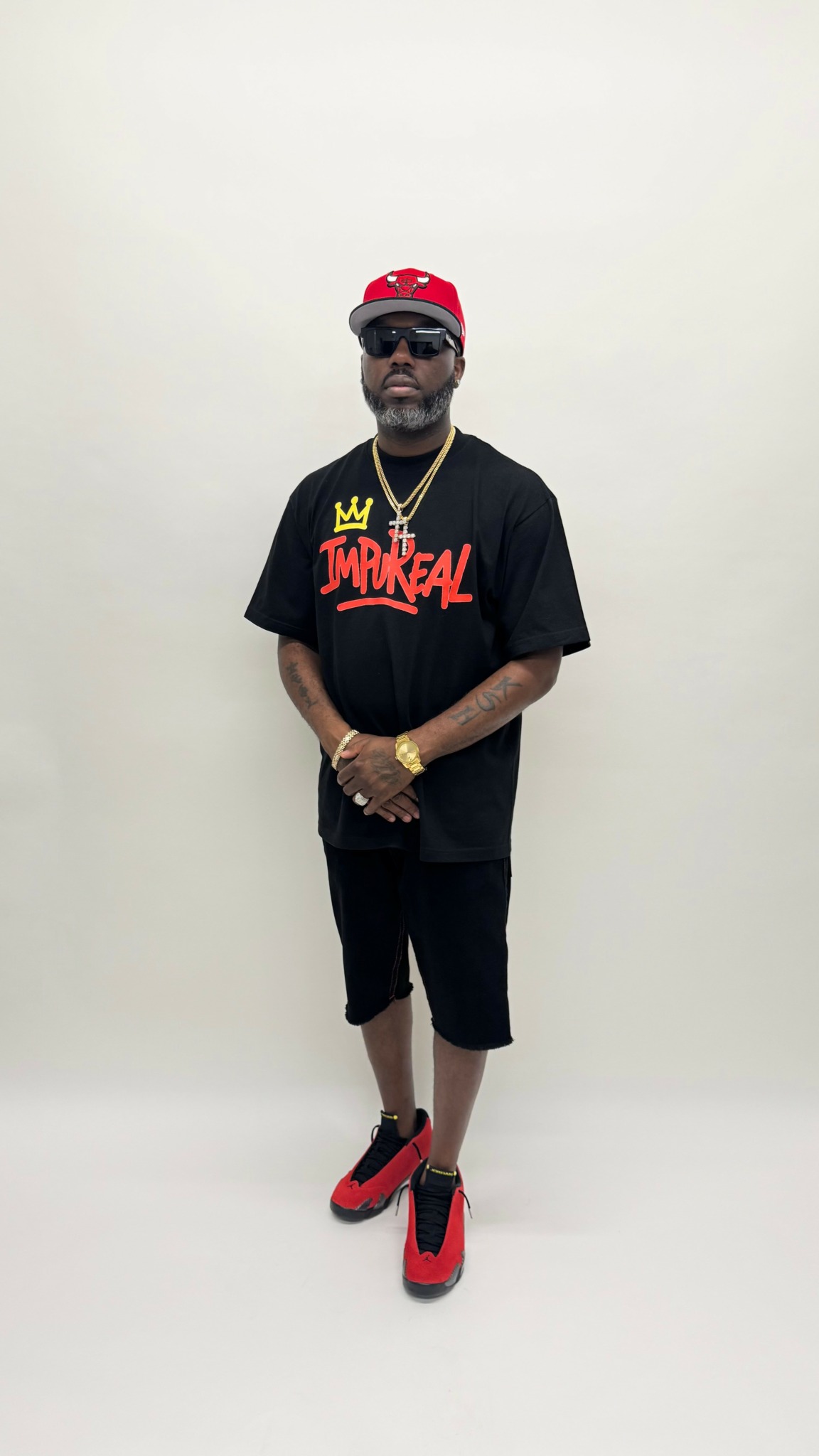 Man standing against a plain white background, wearing sunglasses, a red cap, a black T-shirt with colorful text, black shorts, and red sneakers.