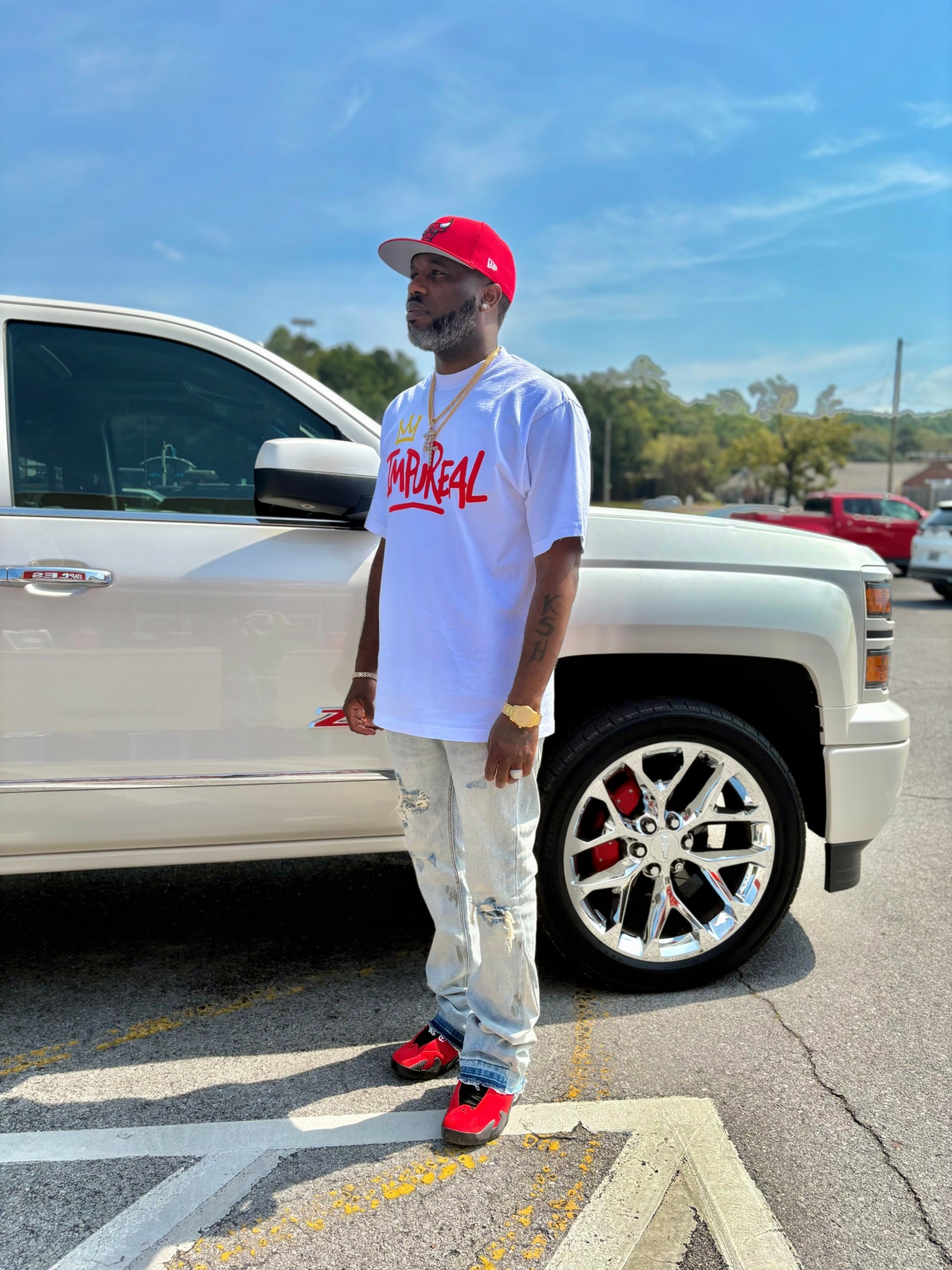 Man wearing a red cap, sunglasses, white t-shirt, and ripped jeans standing next to a white vehicle in a parking lot.