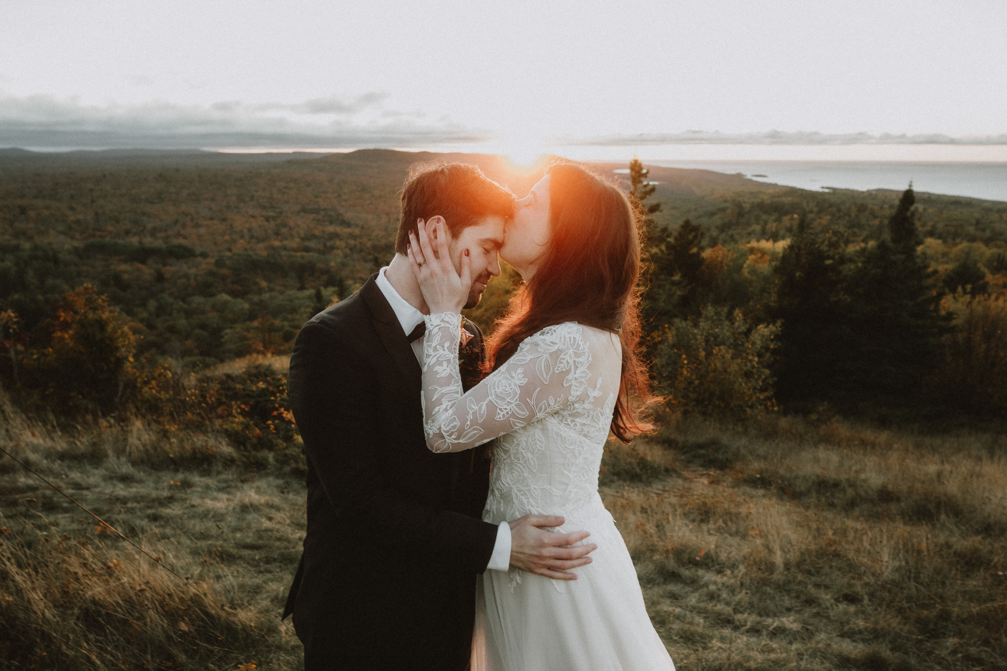 A couple in wedding attire stands close together outdoors at sunset, embracing and touching foreheads, with a landscape background.