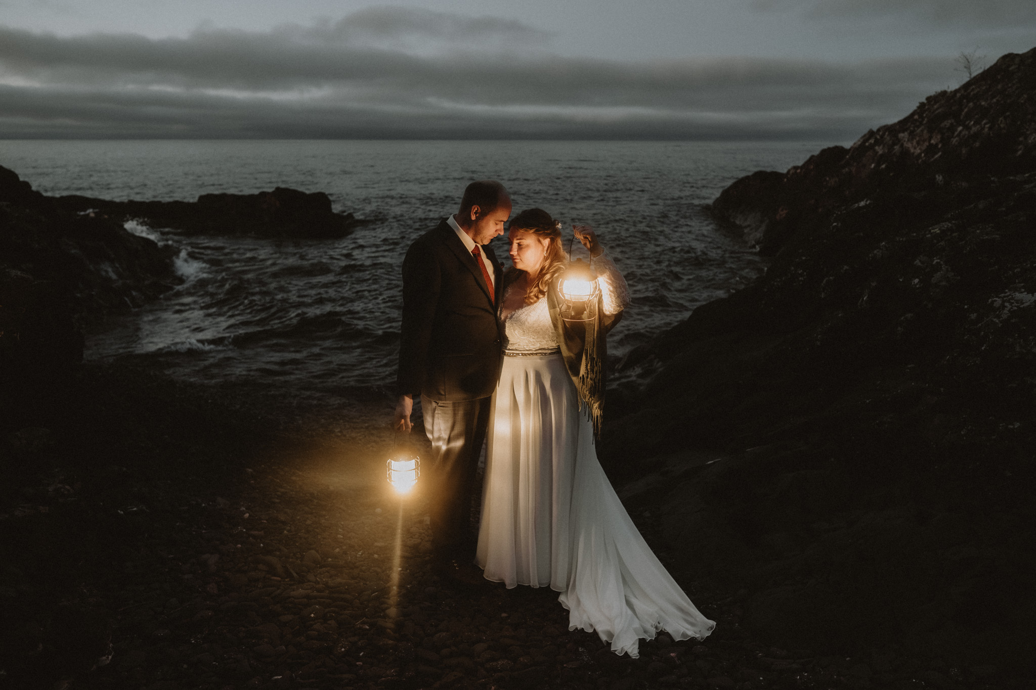 A couple in wedding attire stands on rocks near the ocean at night, holding lanterns with a dark cloudy sky.