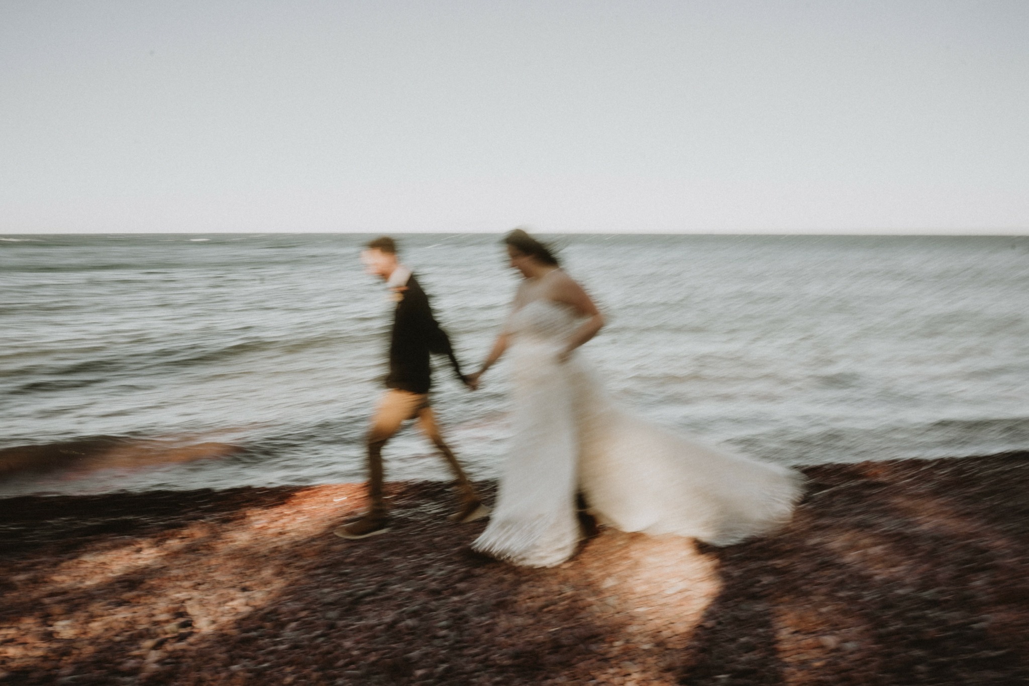A couple holding hands walking on a beach with the ocean in the background, one in a white dress and the other in dark clothing.