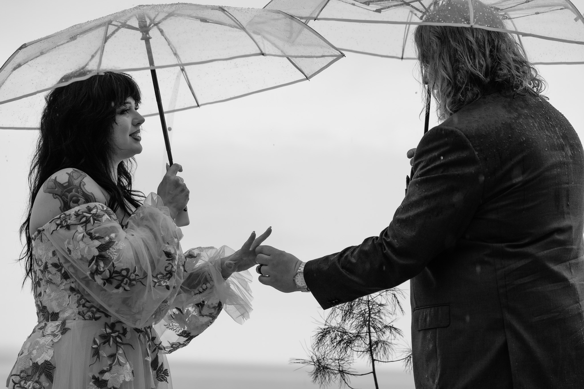 Two people exchange rings under an umbrella outdoors, with a cloudy sky in the background.