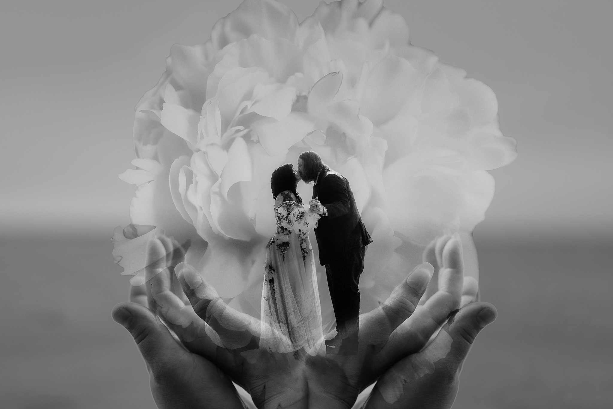 Two people holding a bouquet, with a large flower in the background, and hands in the foreground, in black and white.