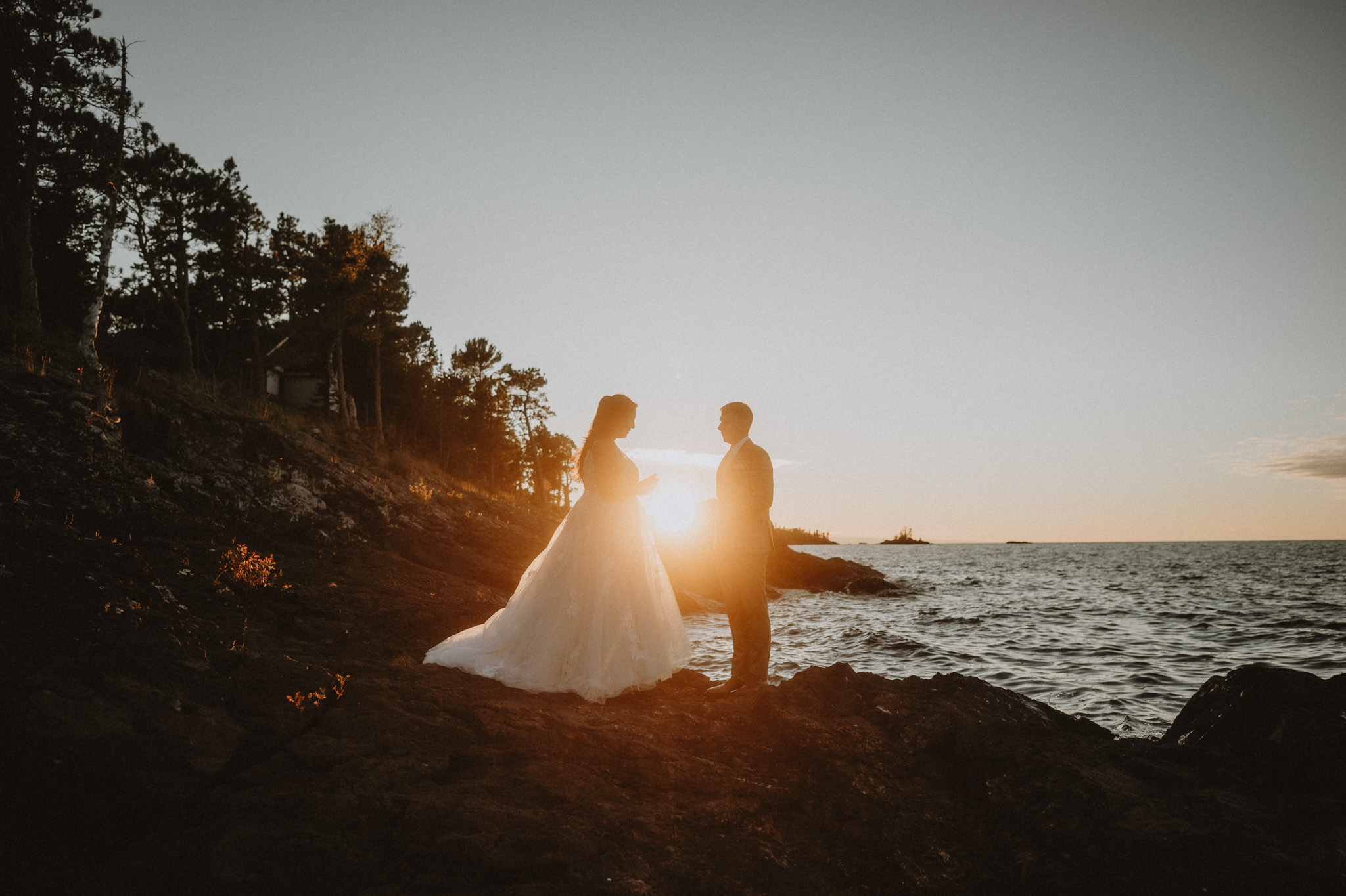 Two people stand on a rocky shoreline at sunset, with the ocean and trees in the background.