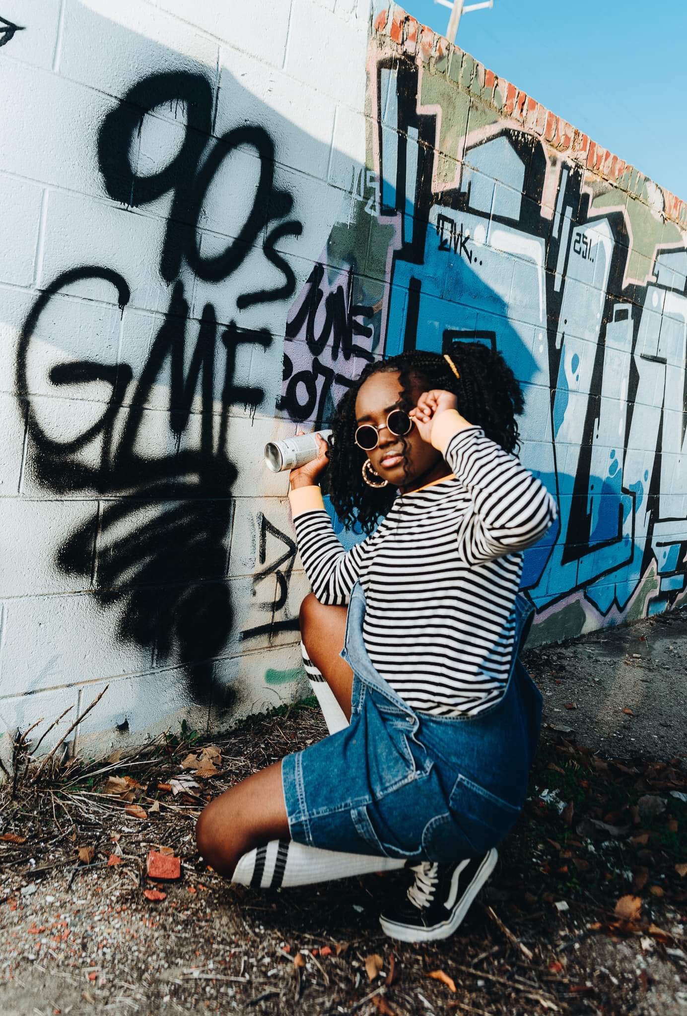 Person crouching in front of graffiti wall, wearing striped shirt, denim shorts, sunglasses, and sneakers, with one hand on head.