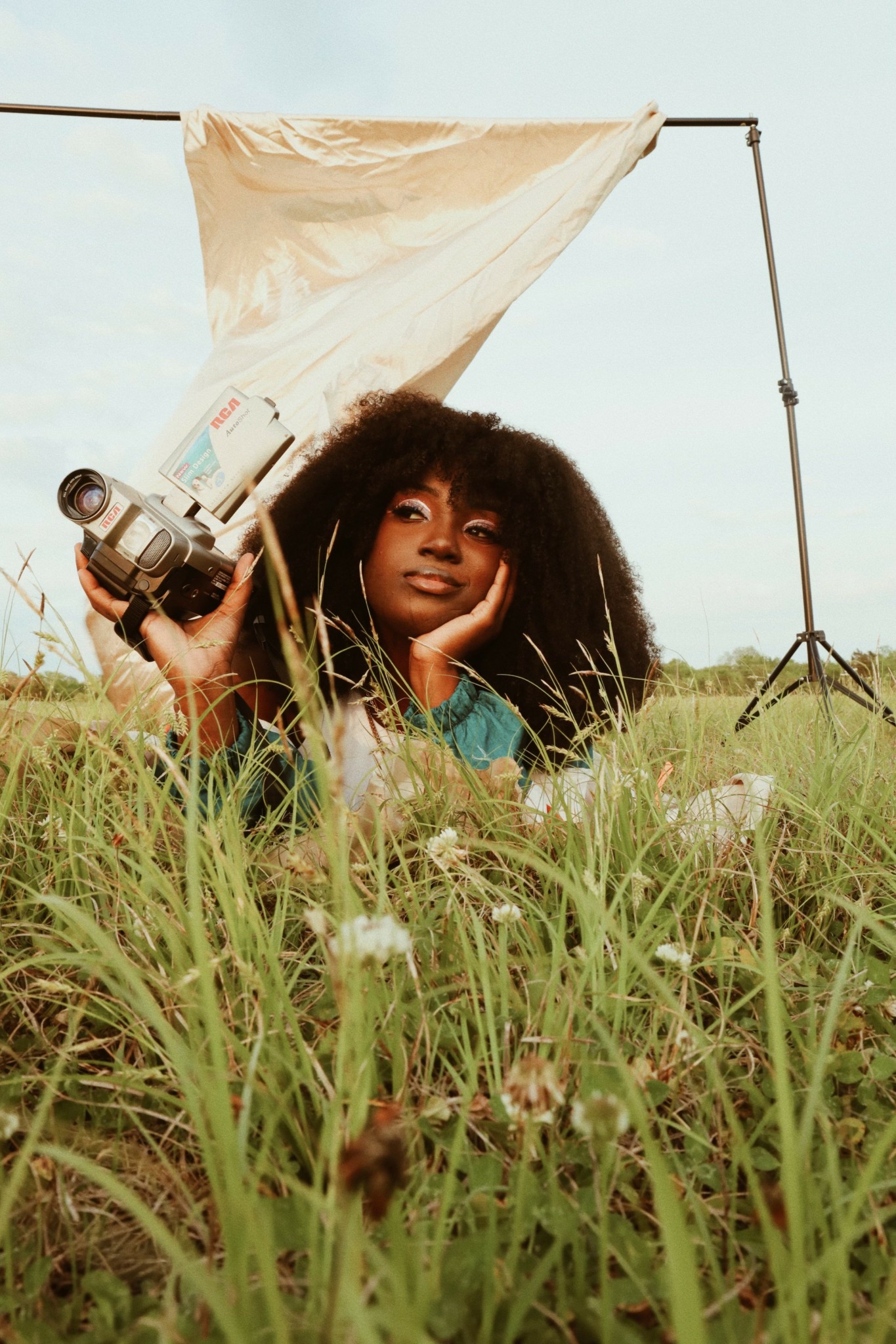 Woman with curly hair lying in grass, holding a camera, with photography equipment and a backdrop behind her.