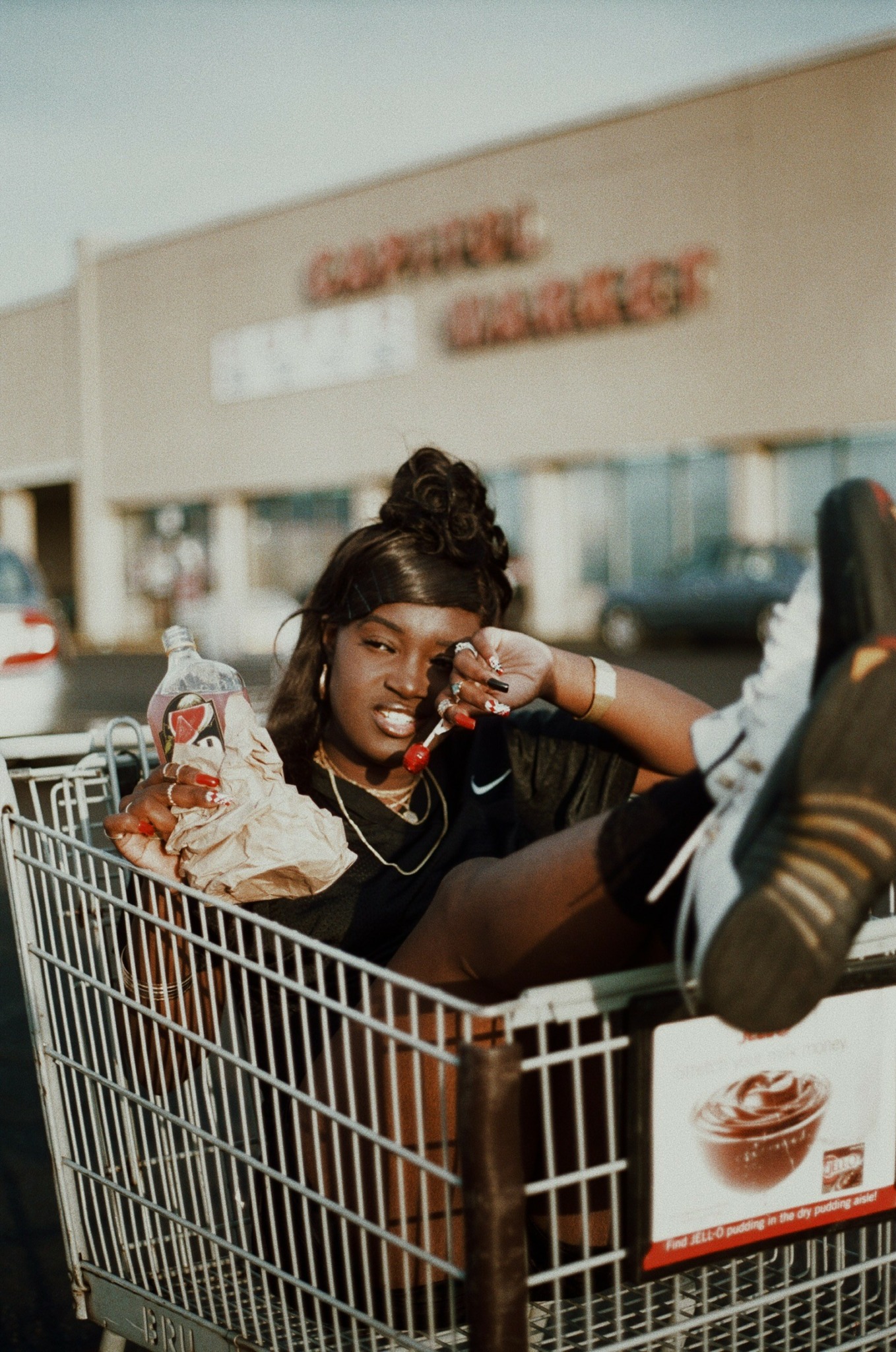 Person sitting in a shopping cart outside a store, with a large building in the background.