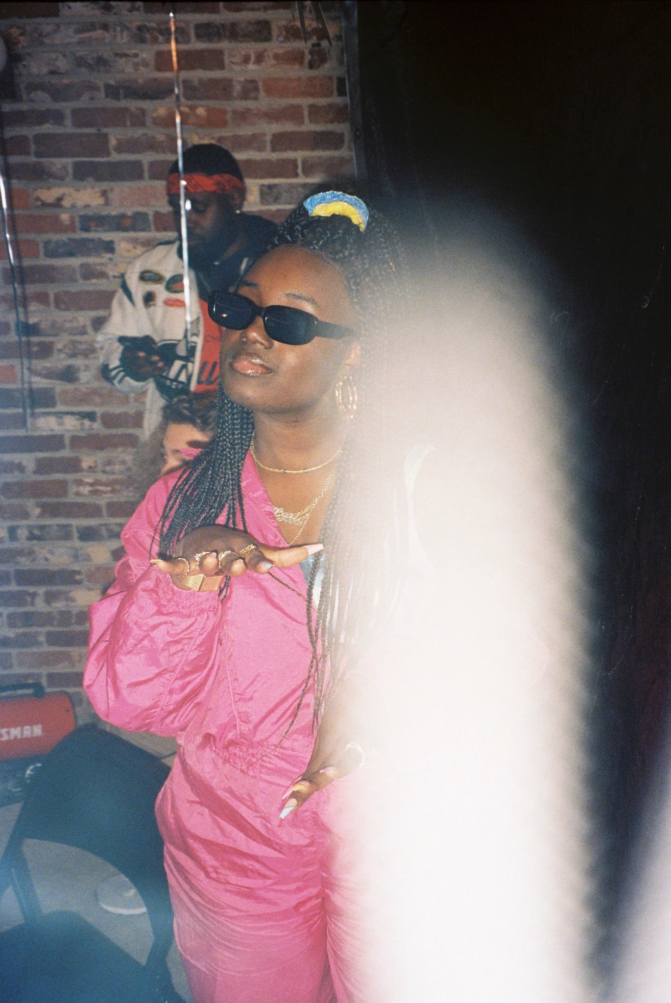 Woman wearing sunglasses and pink outfit, smiling, with a brick wall background, partially obscured by a white object.