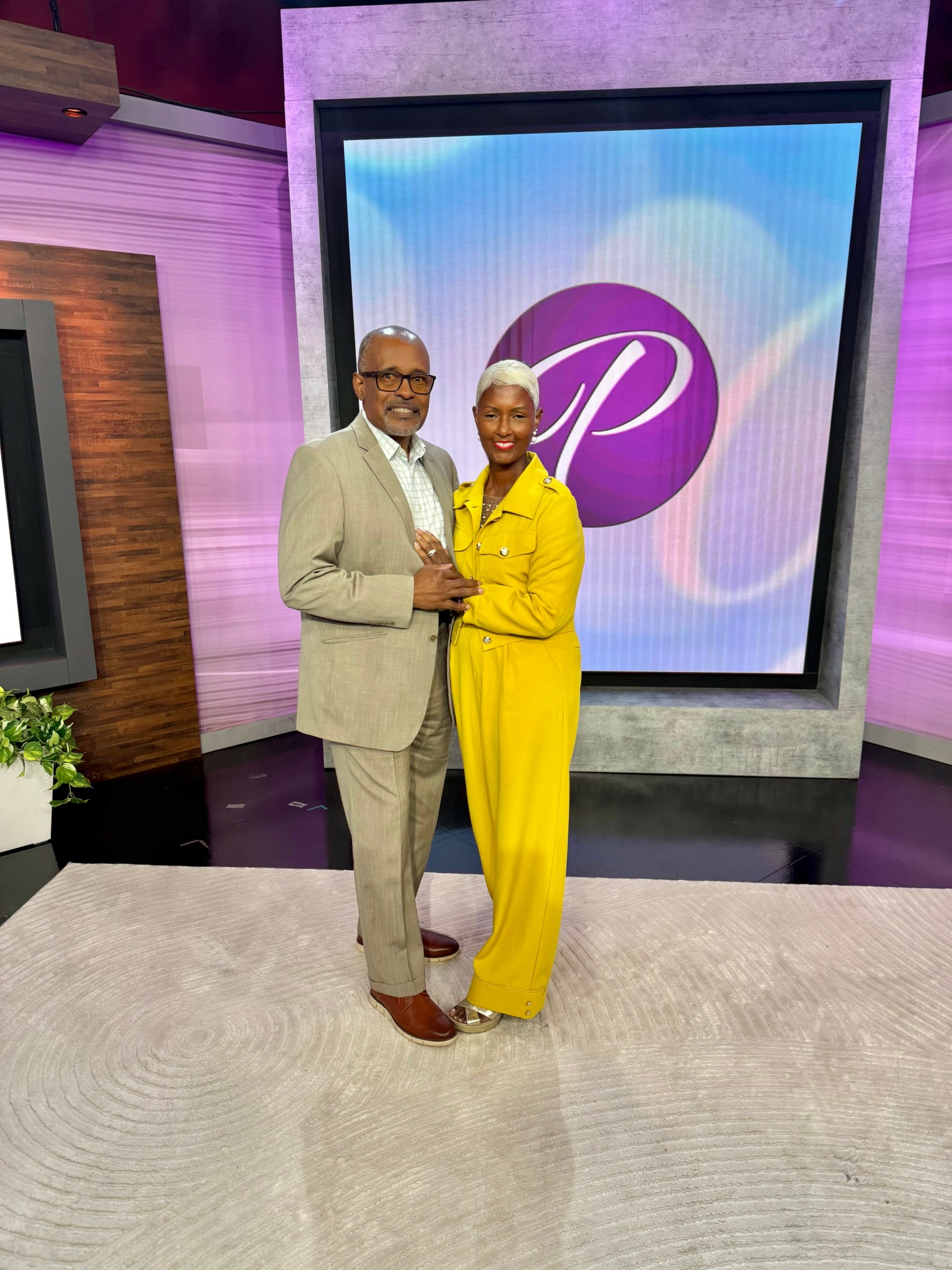Two people, a man in a beige suit and a woman in a yellow outfit, stand together smiling in a studio with a purple and blue background.