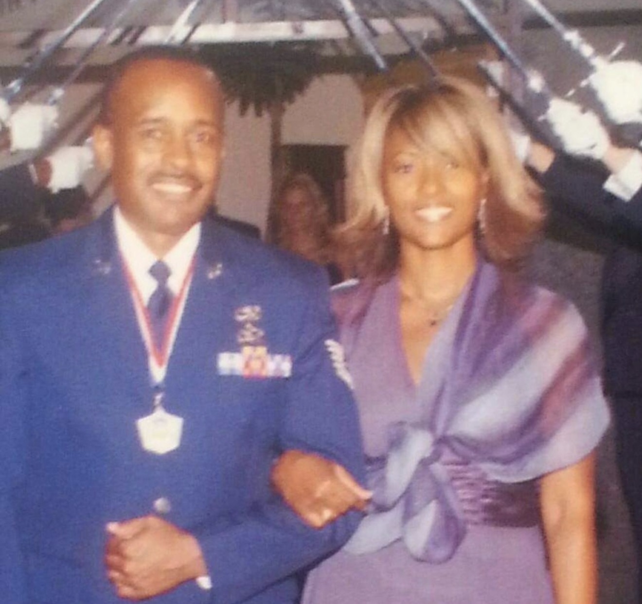 A man in a military uniform and a woman in a dress stand together, smiling, at an event with decorations overhead.
