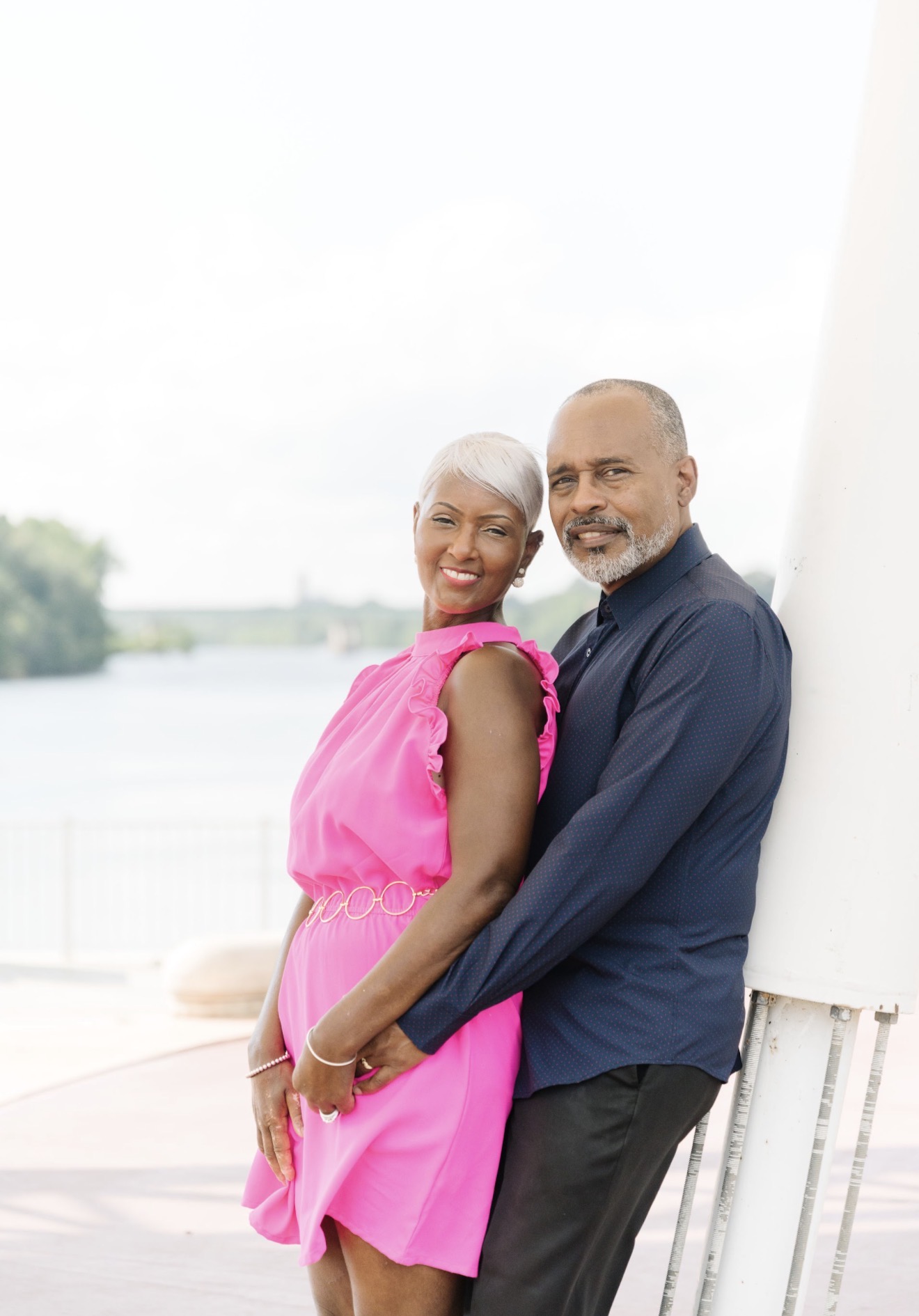 A smiling couple leaning against a white wall outdoors, with water and trees in the background.