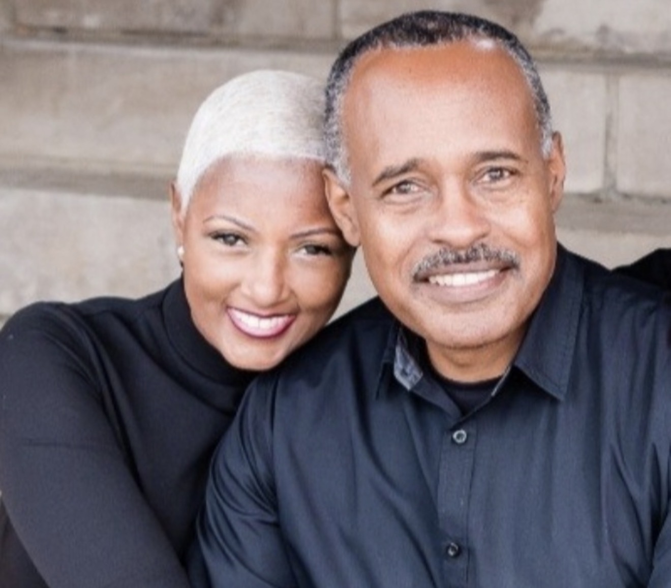 A smiling woman and man taking a selfie outdoors, close together, with a stone wall background.
