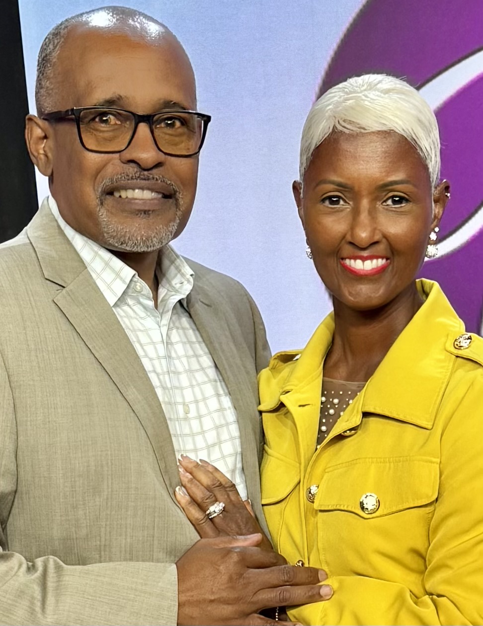 A man and woman stand together, smiling, with the woman wearing a bright yellow jacket and the man in a beige suit.