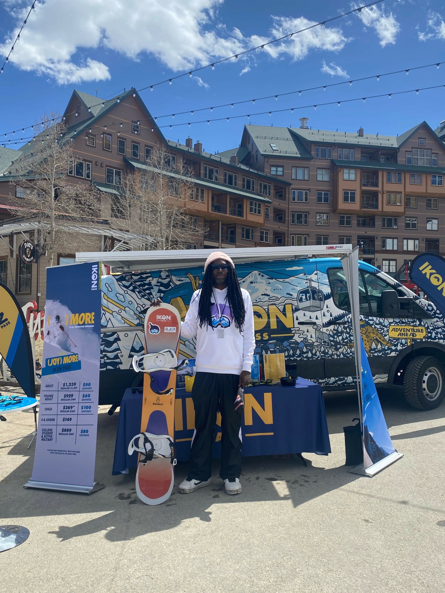 Person standing next to a promotional booth with banners and a table, in an outdoor urban setting with buildings and a blue sky.