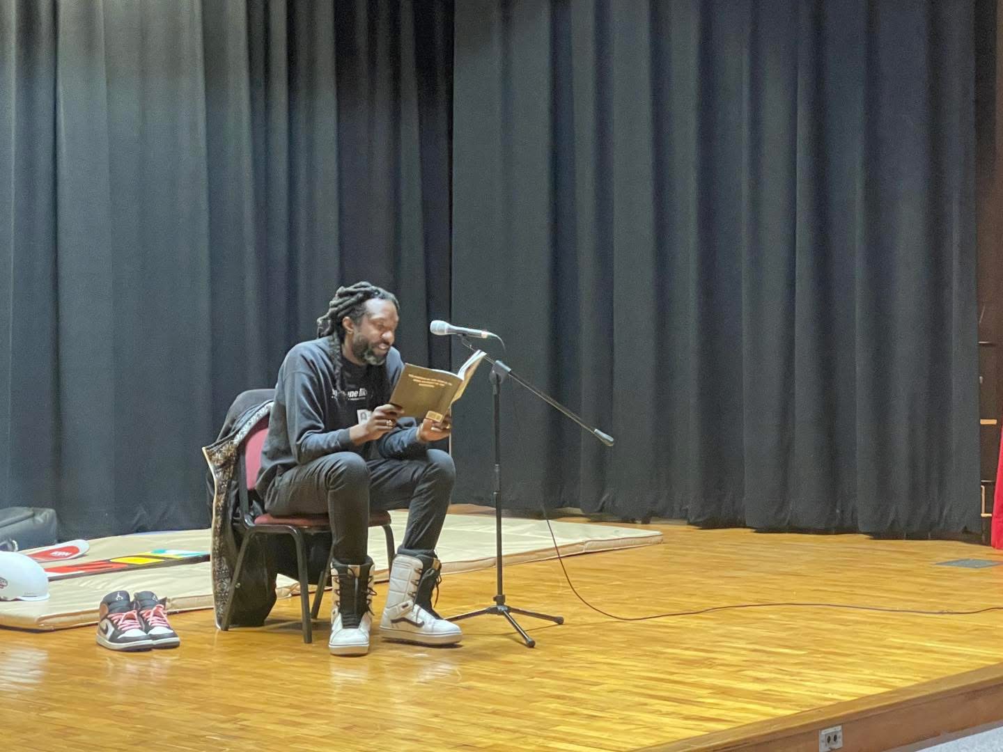 Person sitting on a chair reading a book on stage with a microphone, black curtains in the background, wooden floor.