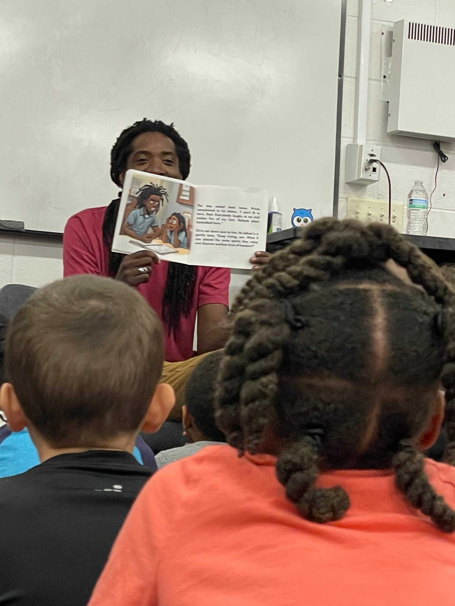 Person reading a book to children in a classroom, with children sitting and listening attentively.