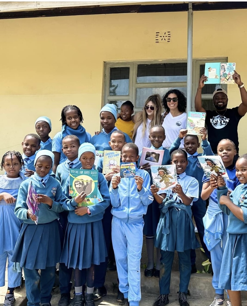 Group of children in blue uniforms and adults standing outside a yellow building, holding books and smiling.