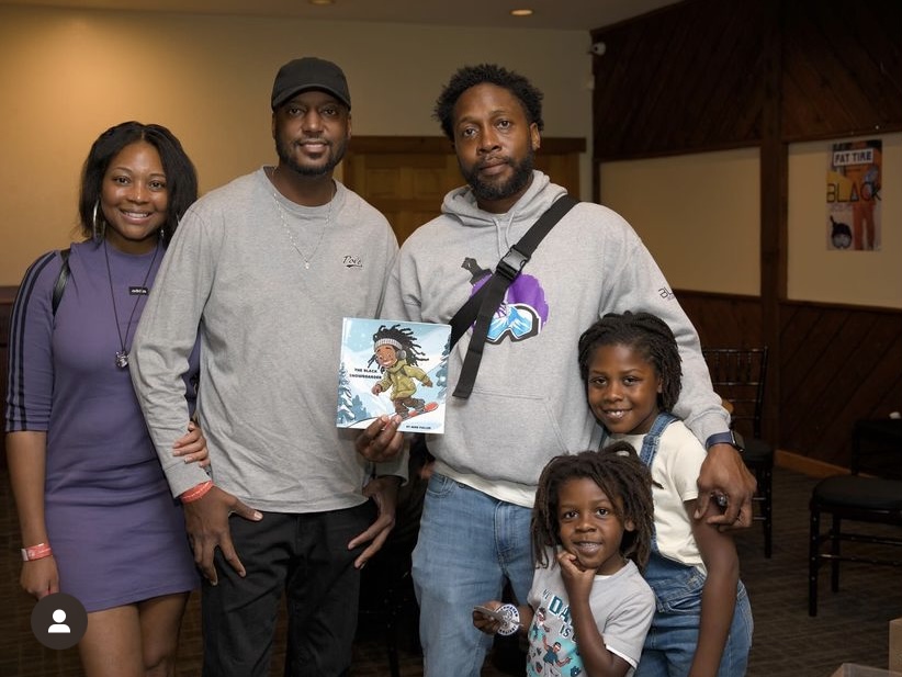 Four people, two adults and two children, standing indoors, smiling, with one holding a colorful book or artwork.
