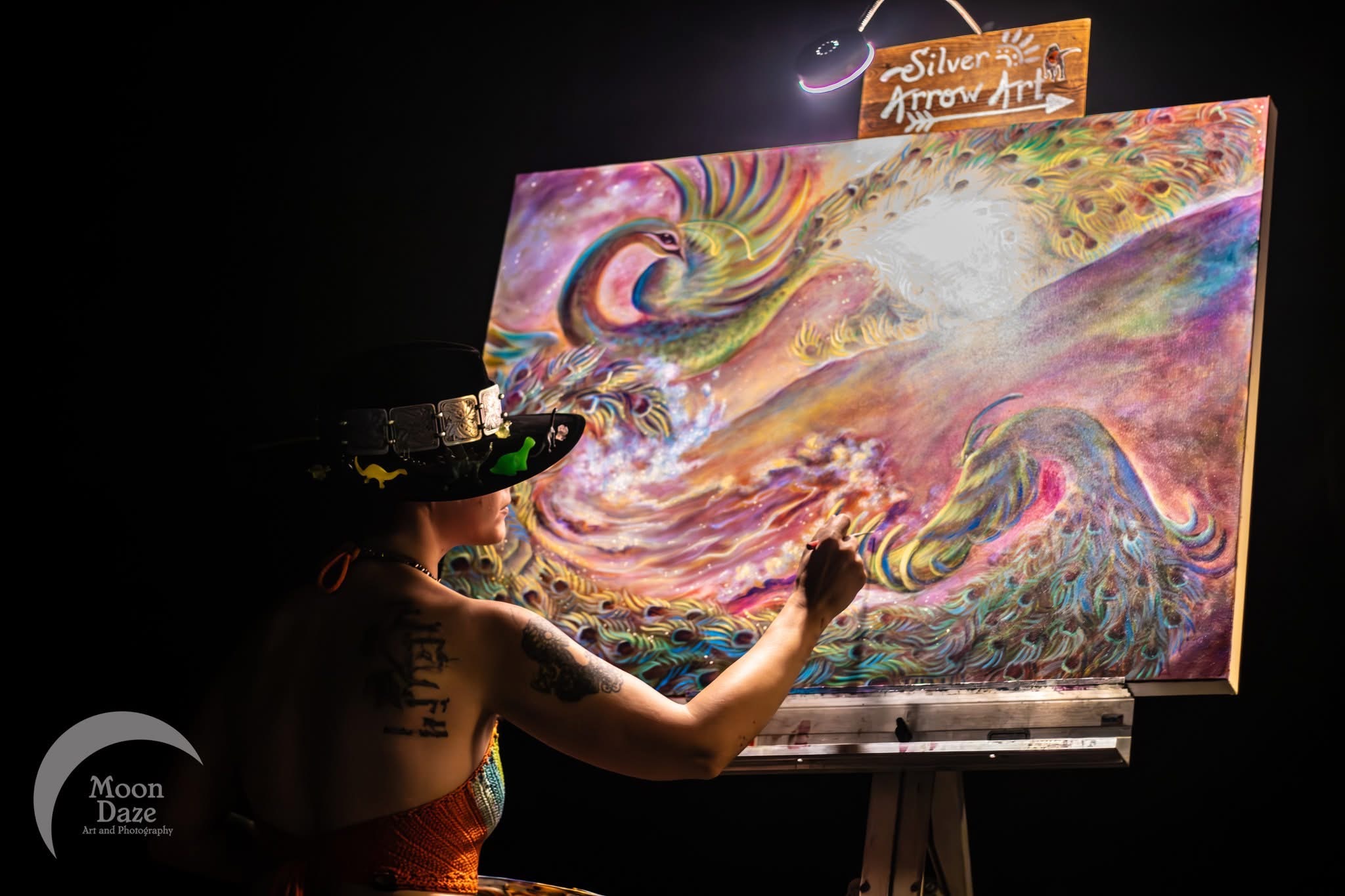 Artist painting a colorful abstract on a large canvas with a dark background, wearing a hat and sleeveless top.