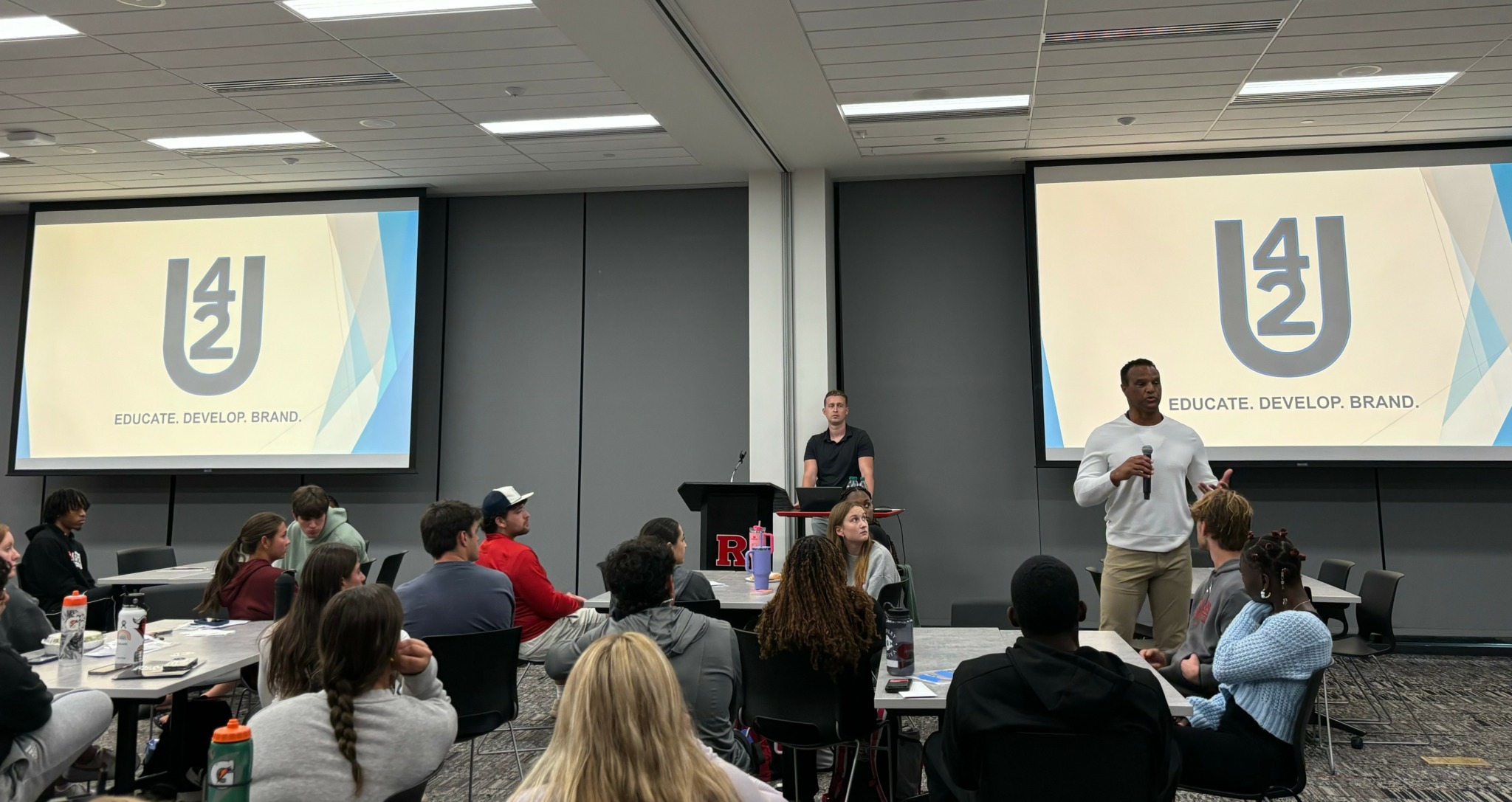 People seated at tables in a conference room, two presenters at the front with screens displaying a logo and text.