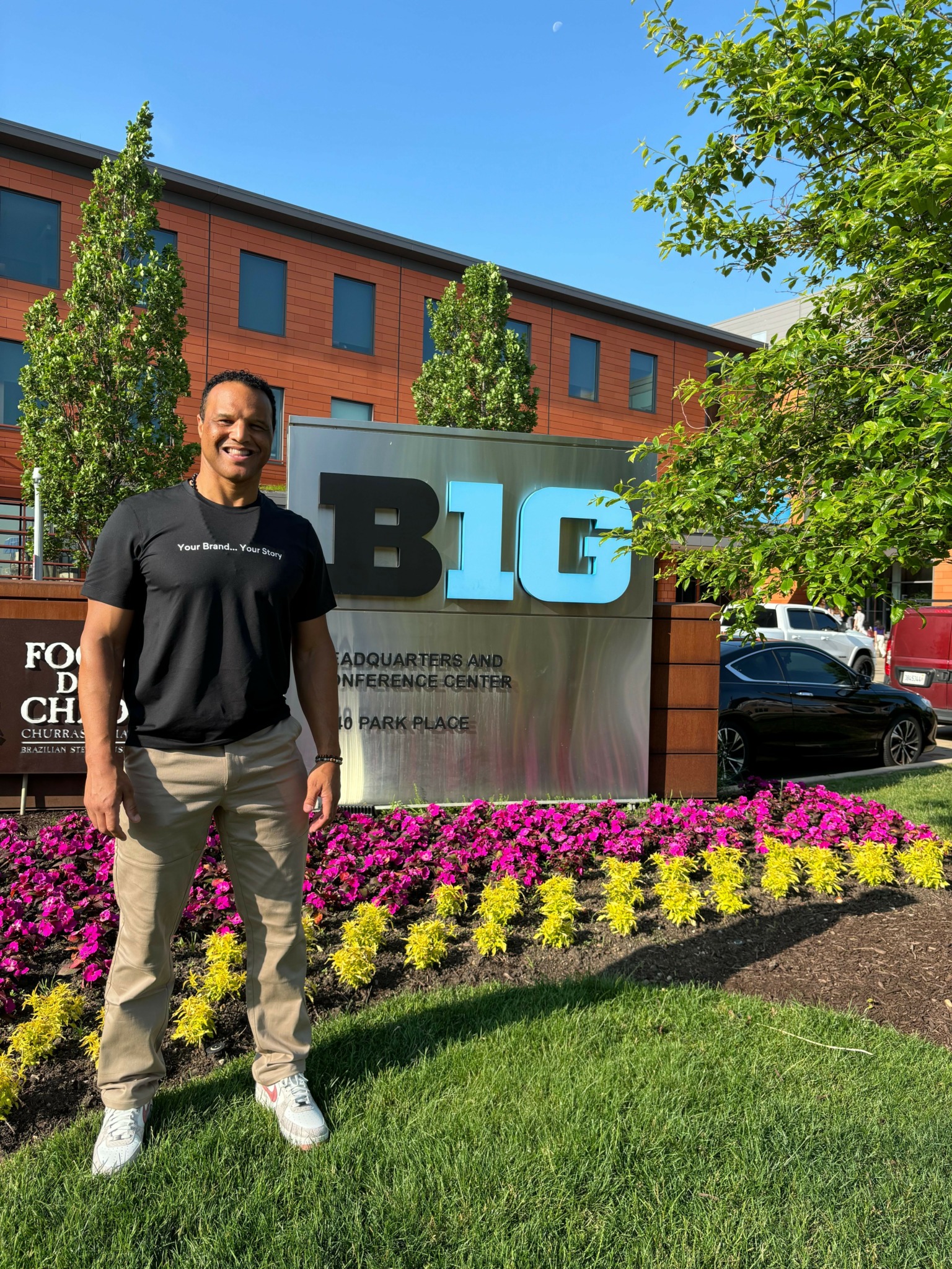 Person standing outdoors near colorful flower bed and a large sign with the letters 'BIG' in front of a brick building.