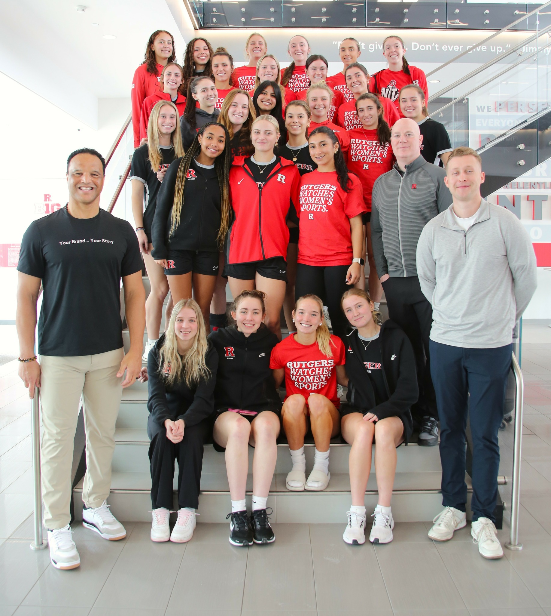 Group of young athletes and coaches on stairs indoors, smiling, wearing sportswear and casual clothing.