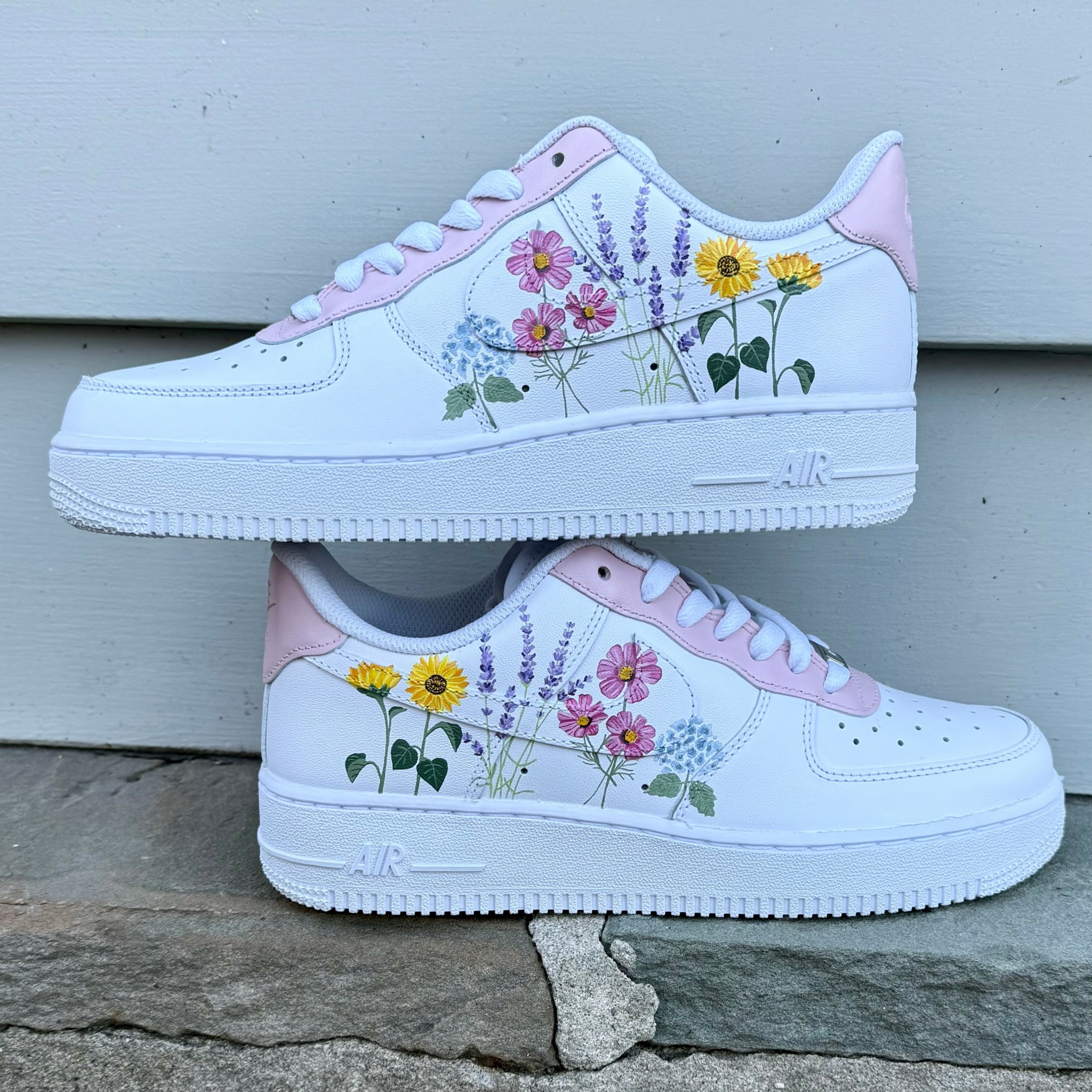 Pair of white sneakers with pink accents and floral illustrations of yellow and pink flowers, placed outdoors on a stone surface.