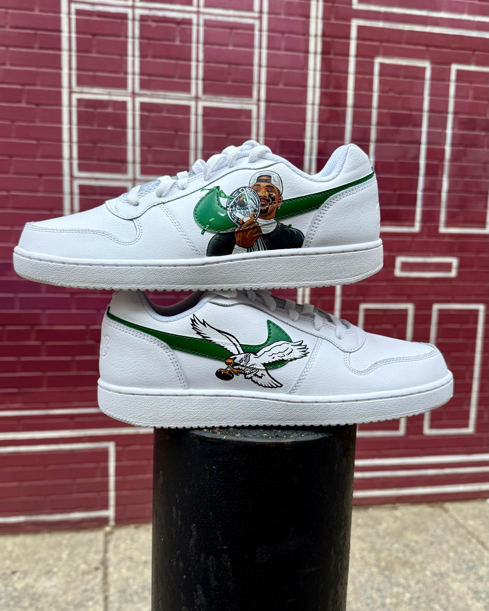 Pair of white sneakers with colorful cartoon character and bird illustrations, displayed against a red brick wall background.