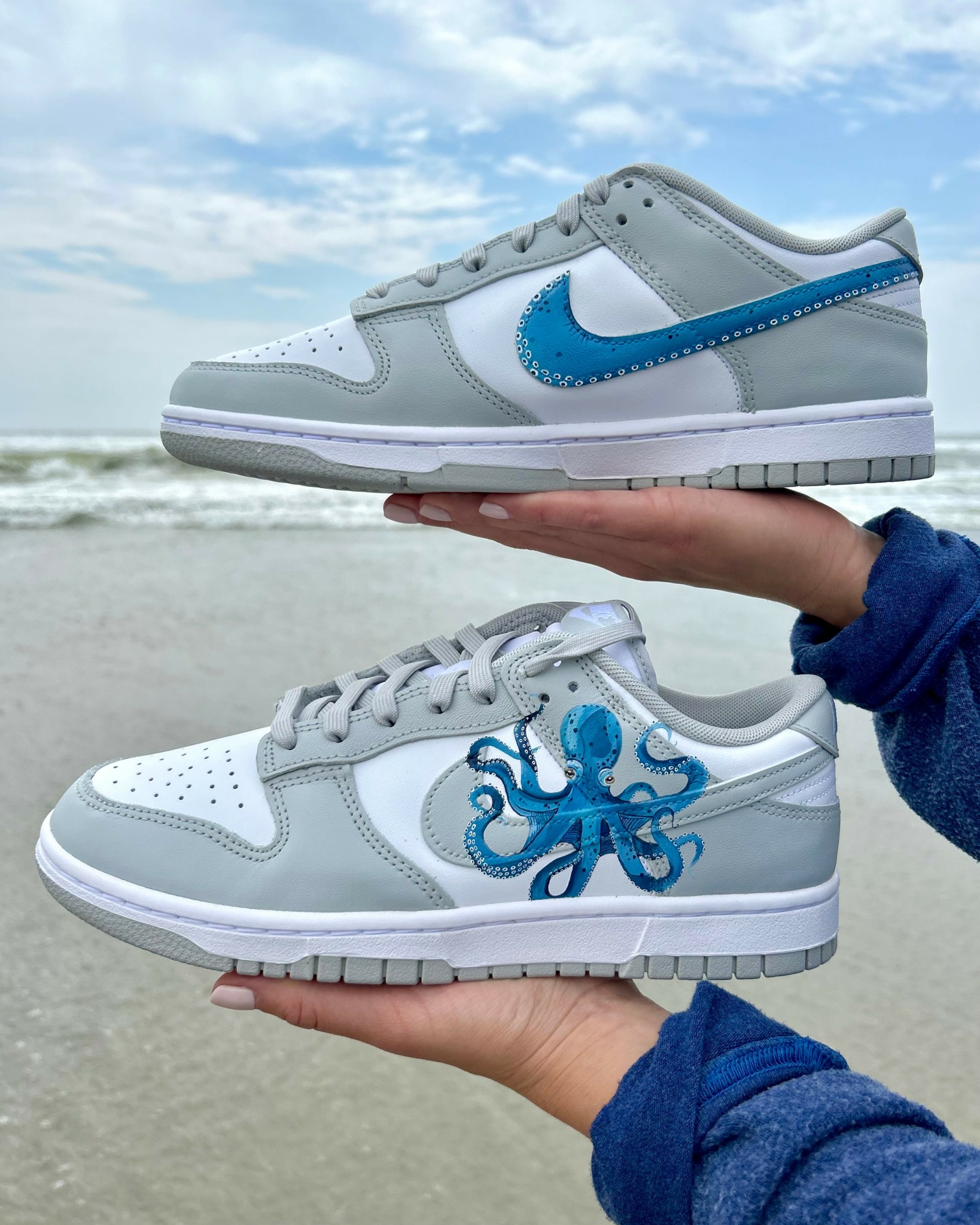 Two sneakers held by hands against a beach background, one with a blue swoosh and the other with a blue octopus design.