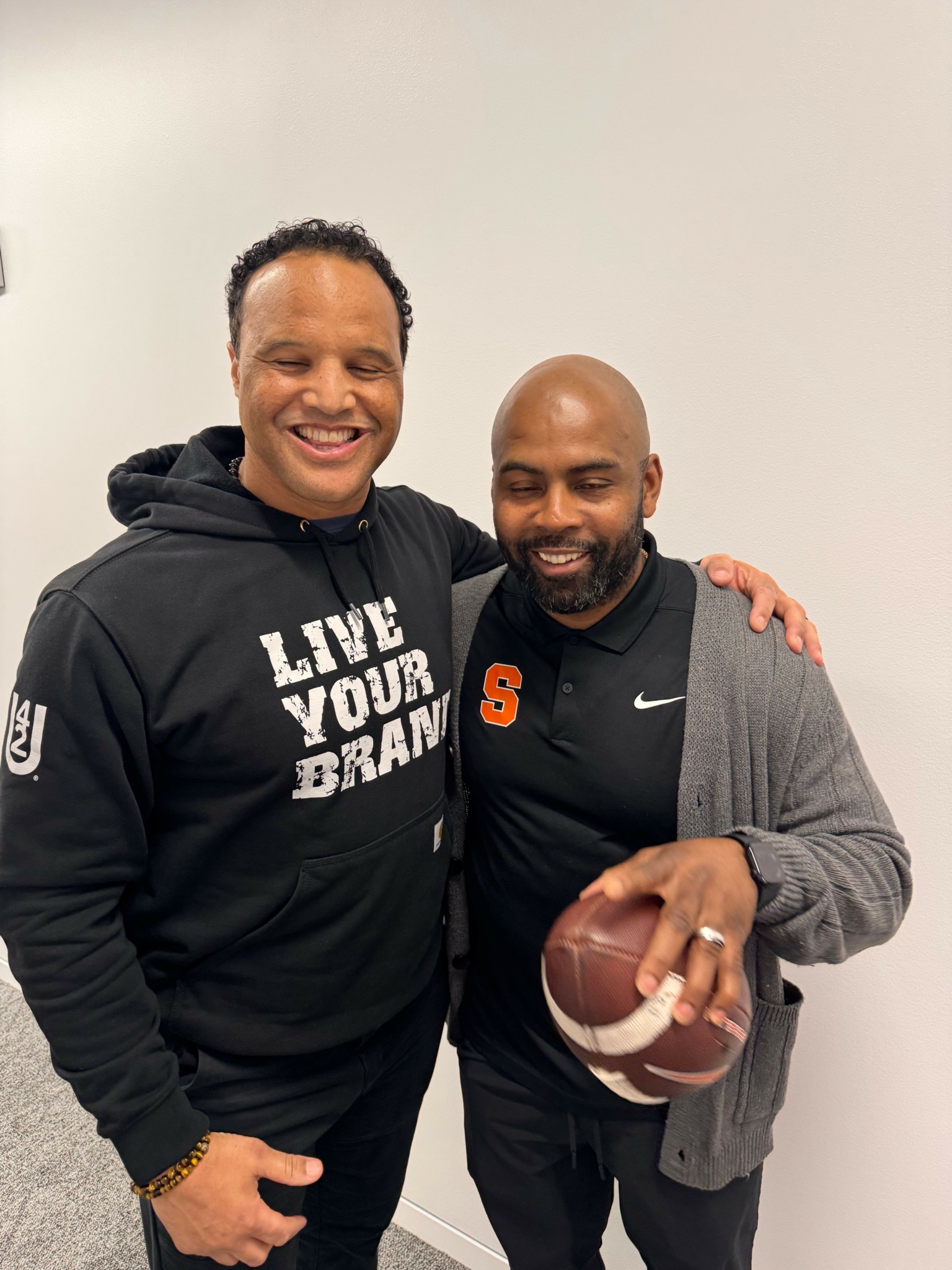 Two men standing together, one holding a football, smiling, against a plain background.