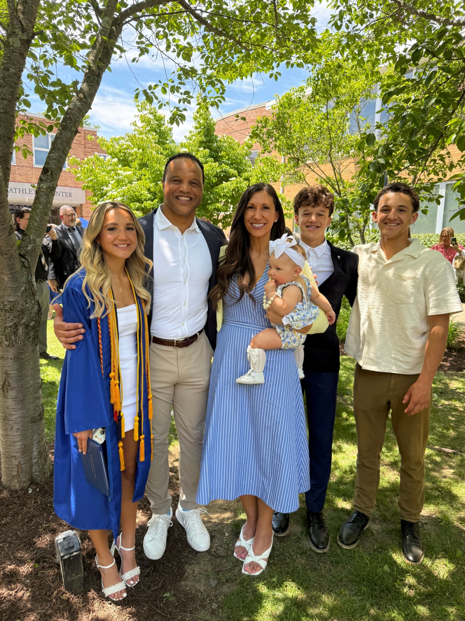 Group of five people outdoors in a park, smiling, with trees and a building in the background.