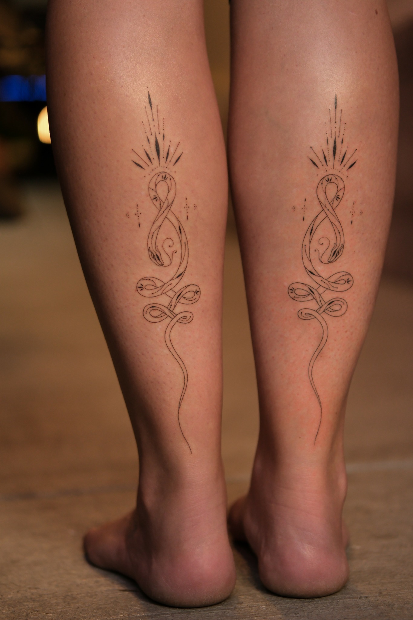 Two legs with matching tattoo designs of a stylized snake intertwined with a vertical line, with radiating lines above the head.
