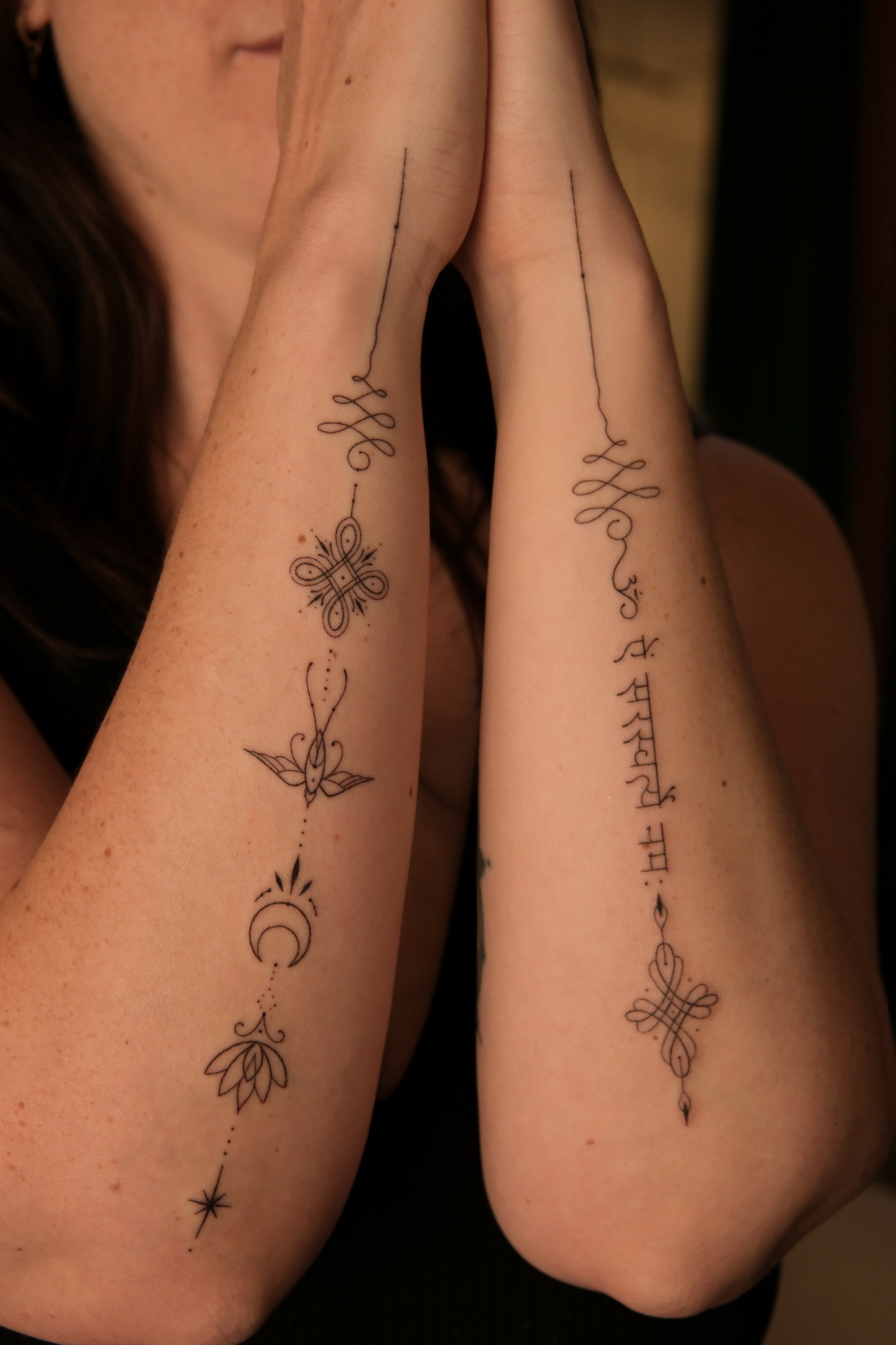 Two arms with tattoos, one with a flower and moon, the other with script and floral designs, raised with palms together.