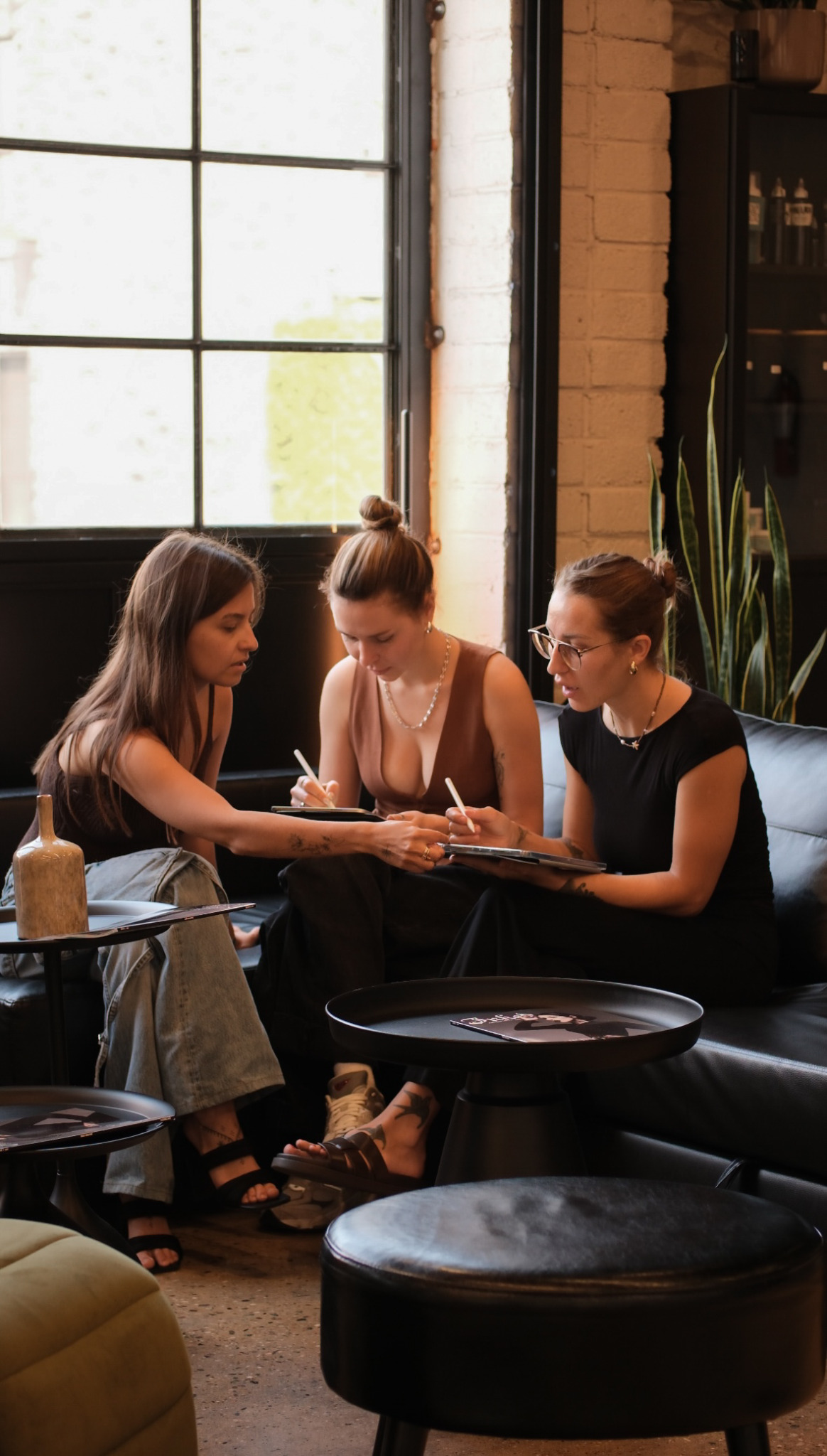 Three women sit on a black sofa, engaged in conversation inside a room with large window and brick wall.