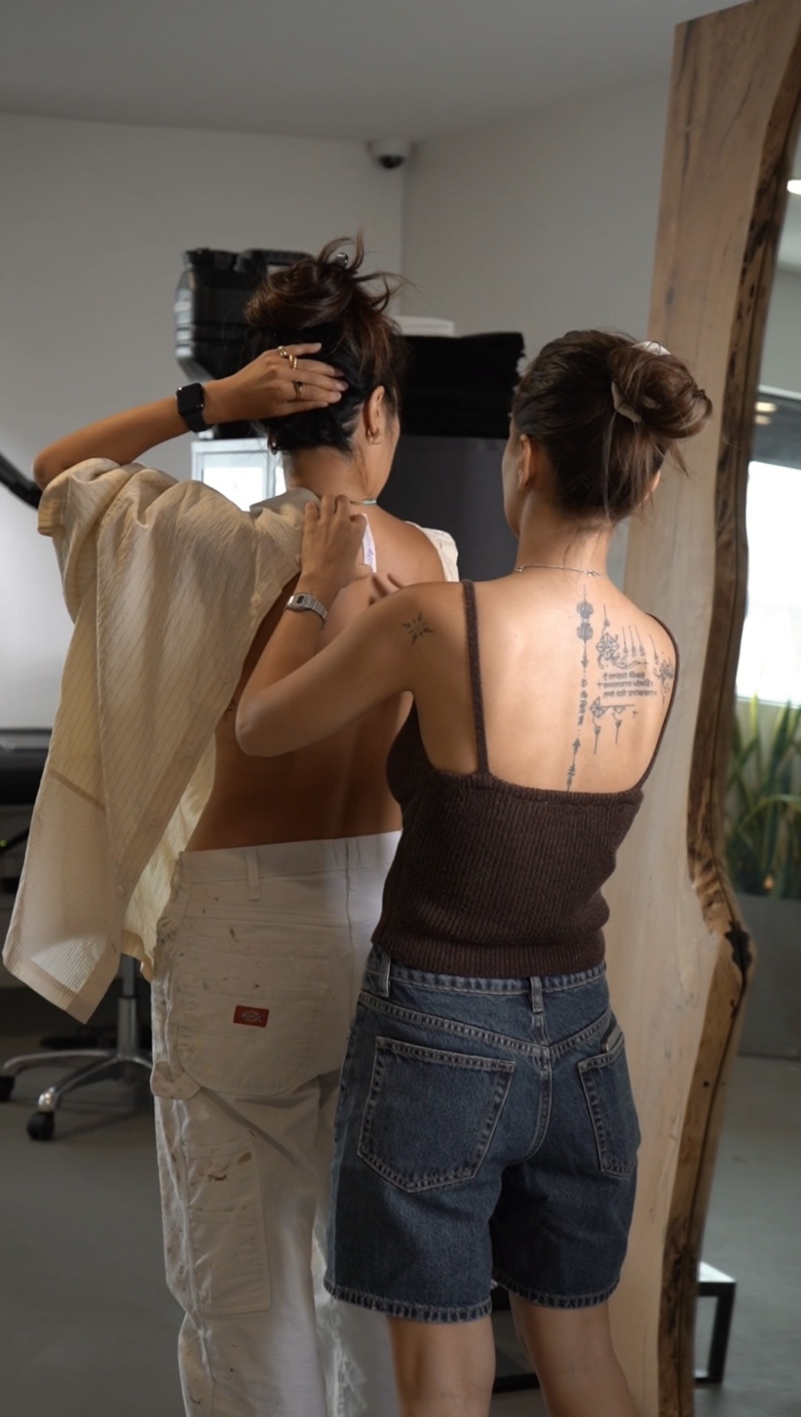 Two women with updo hairstyles stand in front of a mirror, one adjusting her hair, in a room with a plant and furniture.