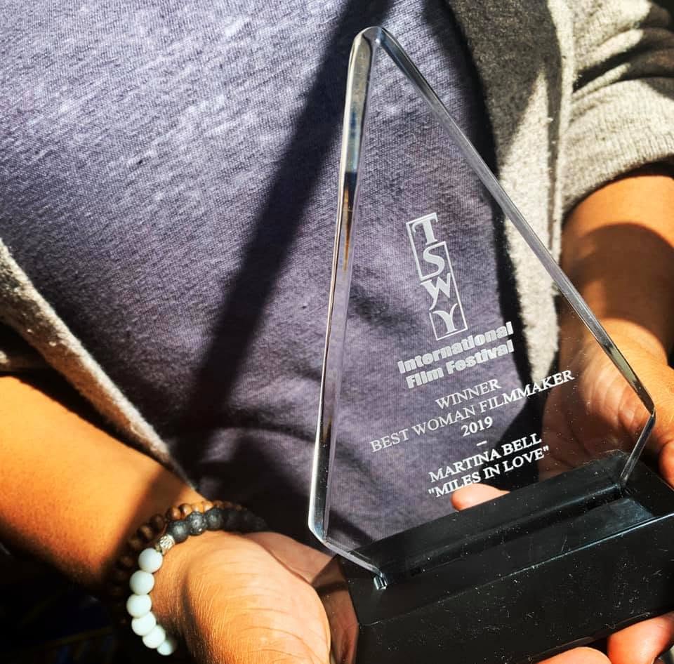 Person holding a glass award with text about an international film festival, wearing a gray shirt and beaded bracelet.