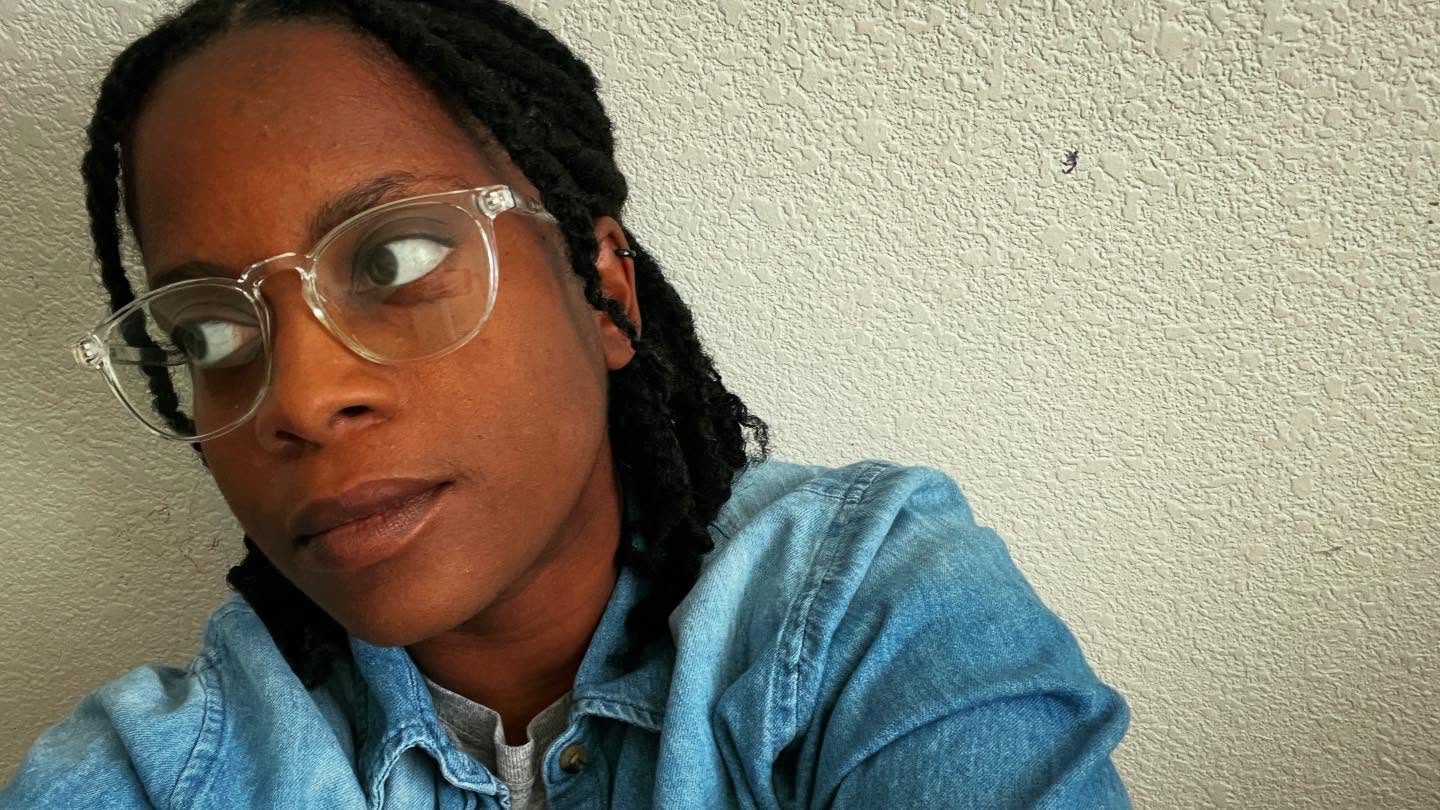 Person with glasses and braided hair wearing a denim jacket, looking to the side, against a plain wall.