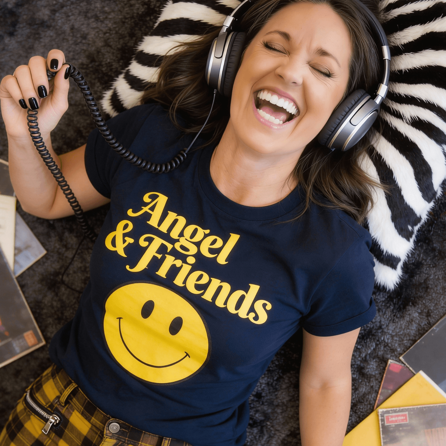 Woman lying on a striped pillow, smiling with headphones, wearing a navy T-shirt with yellow text and smiley face, plaid pants.