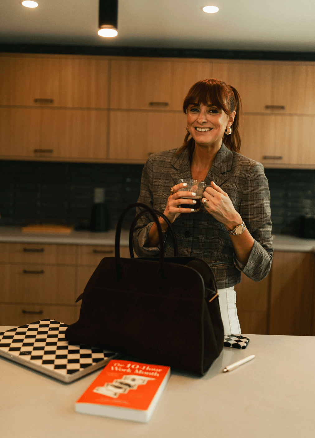 Woman smiling, holding a glass, standing in a kitchen with wooden cabinets, black backsplash, and a white countertop.