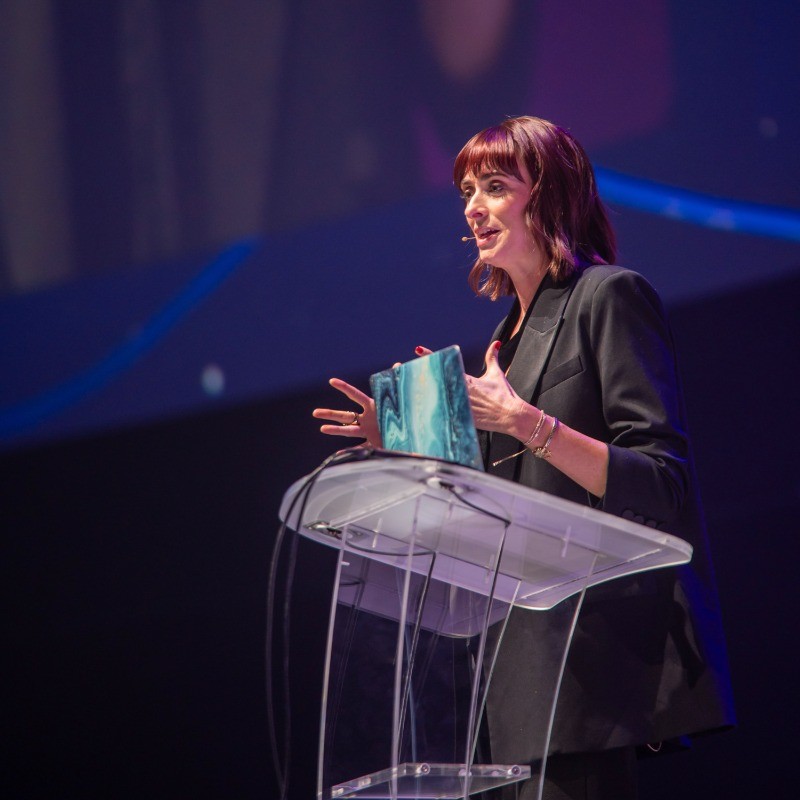 Woman speaking at a transparent podium with a blue gift bag, wearing a dark blazer, in a dark stage setting.