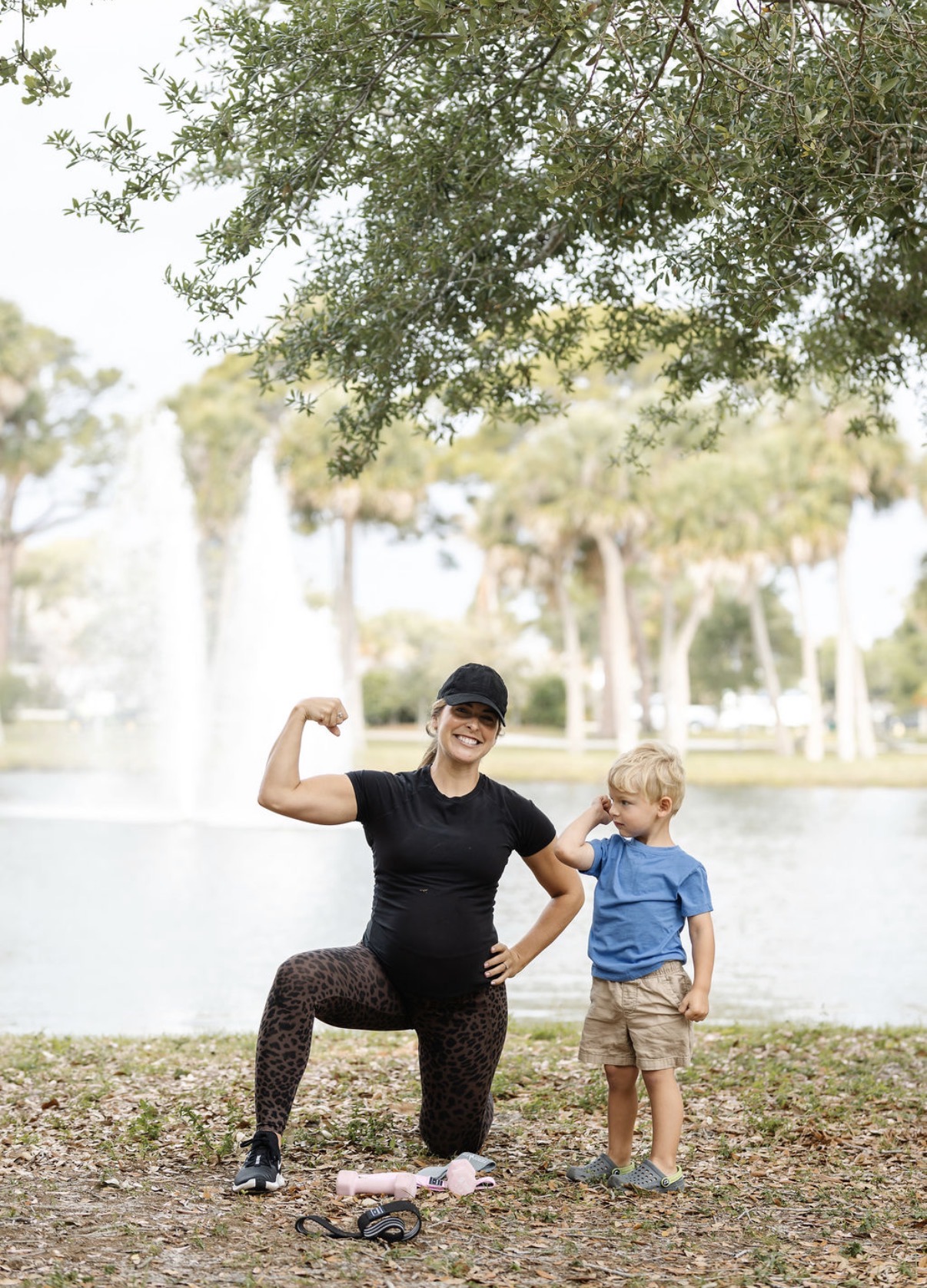 Woman and young boy outdoors near a lake with trees, woman flexing arm, boy looking at her, sunny day.