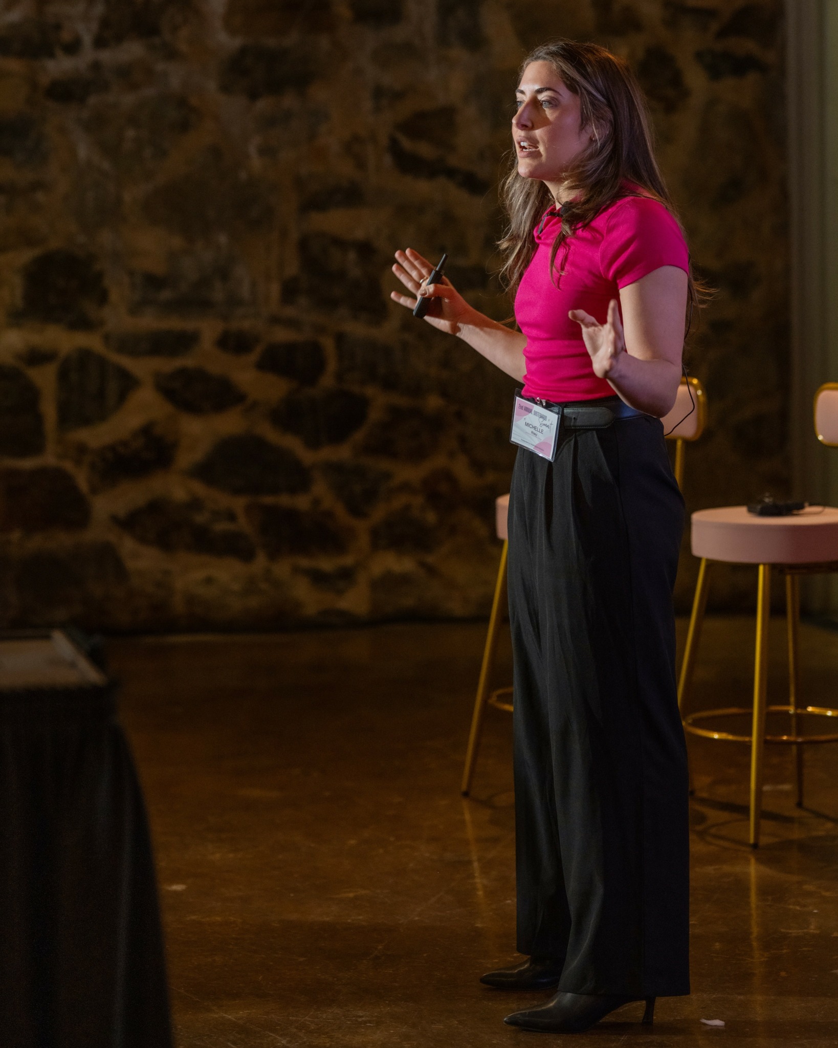 Woman in pink shirt speaking on stage with a stone wall background, gesturing with hands, standing near a chair.